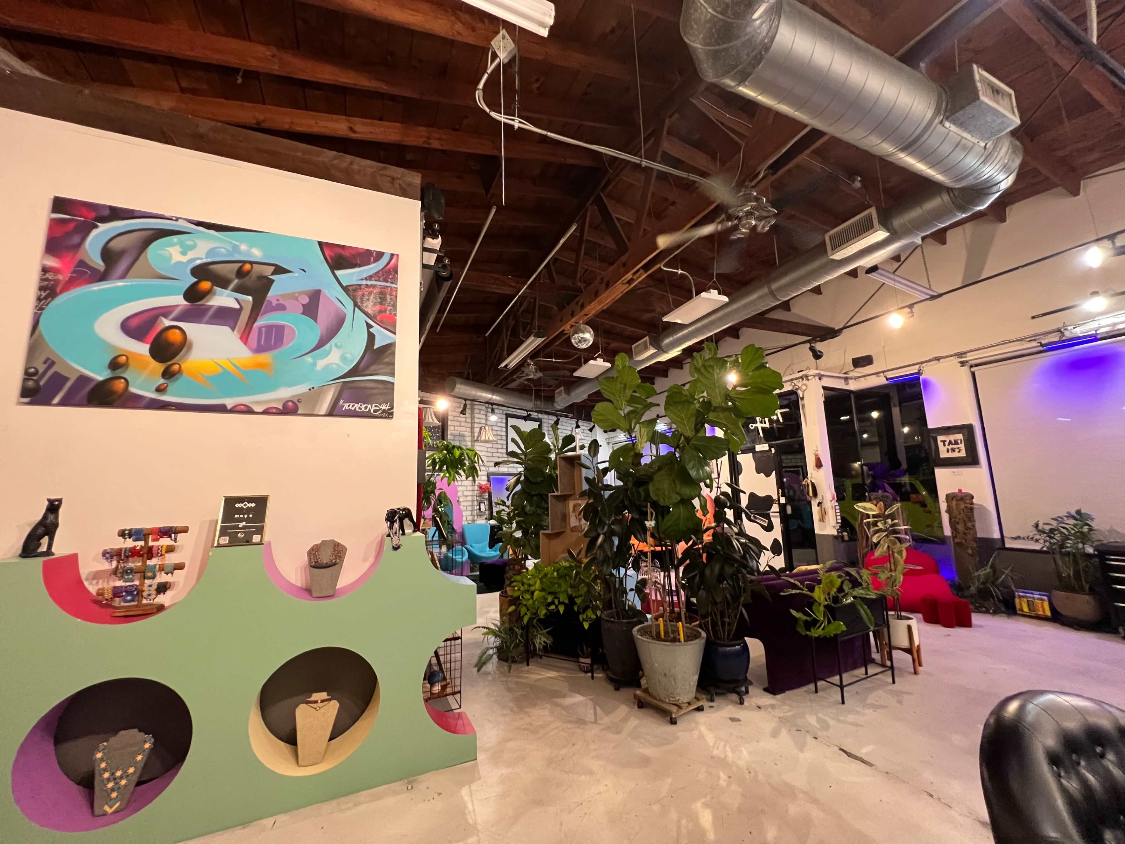 The image shows a modern indoor space with vibrant decor, including plants, colorful furniture, and a mural on the wall.