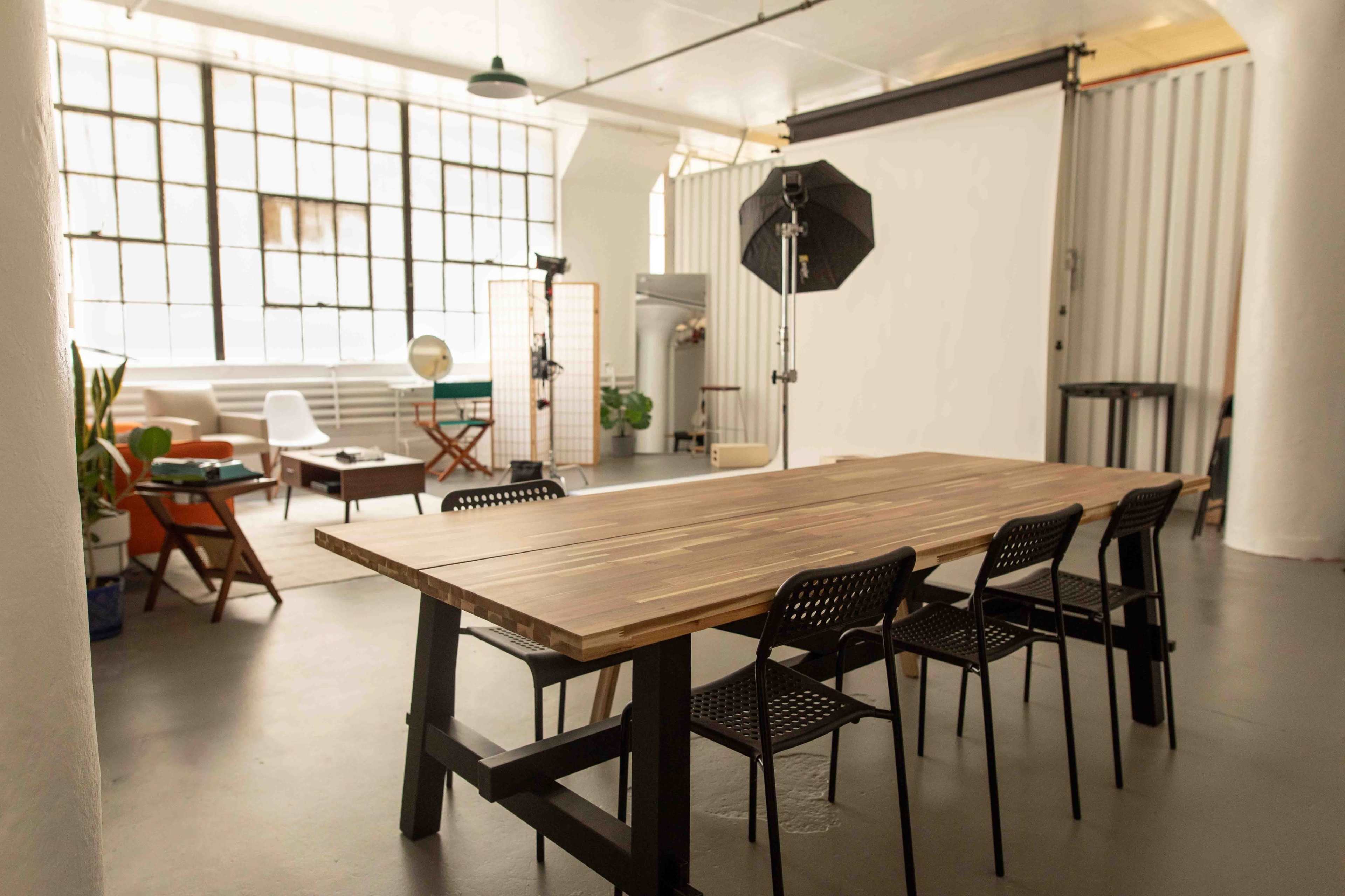 A spacious, well-lit room featuring a large wooden table surrounded by chairs, with a backdrop area and various studio equipment in the corner.