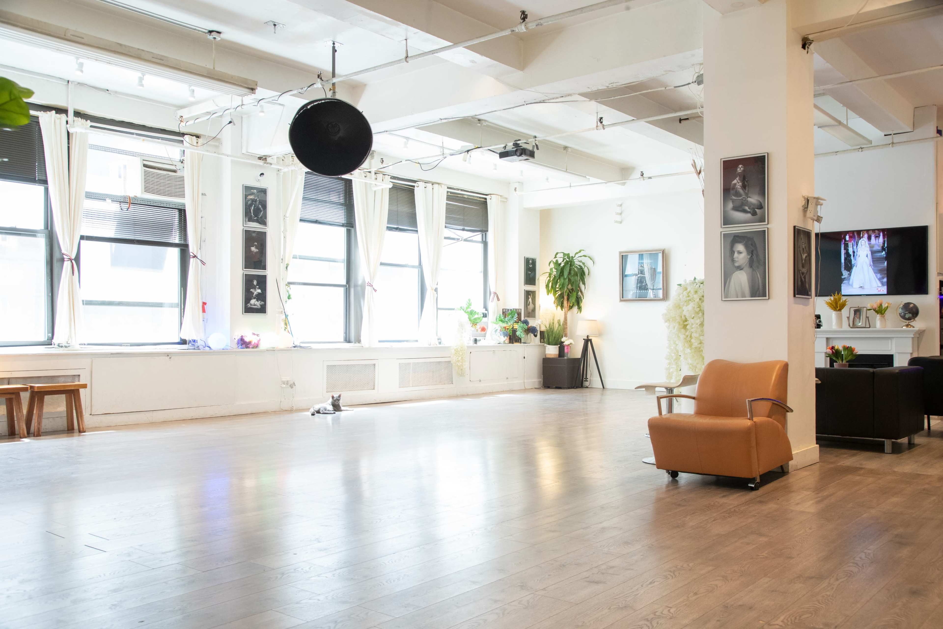 The image shows a well-lit studio space with large windows, wooden flooring, and minimal furniture including an orange chair.