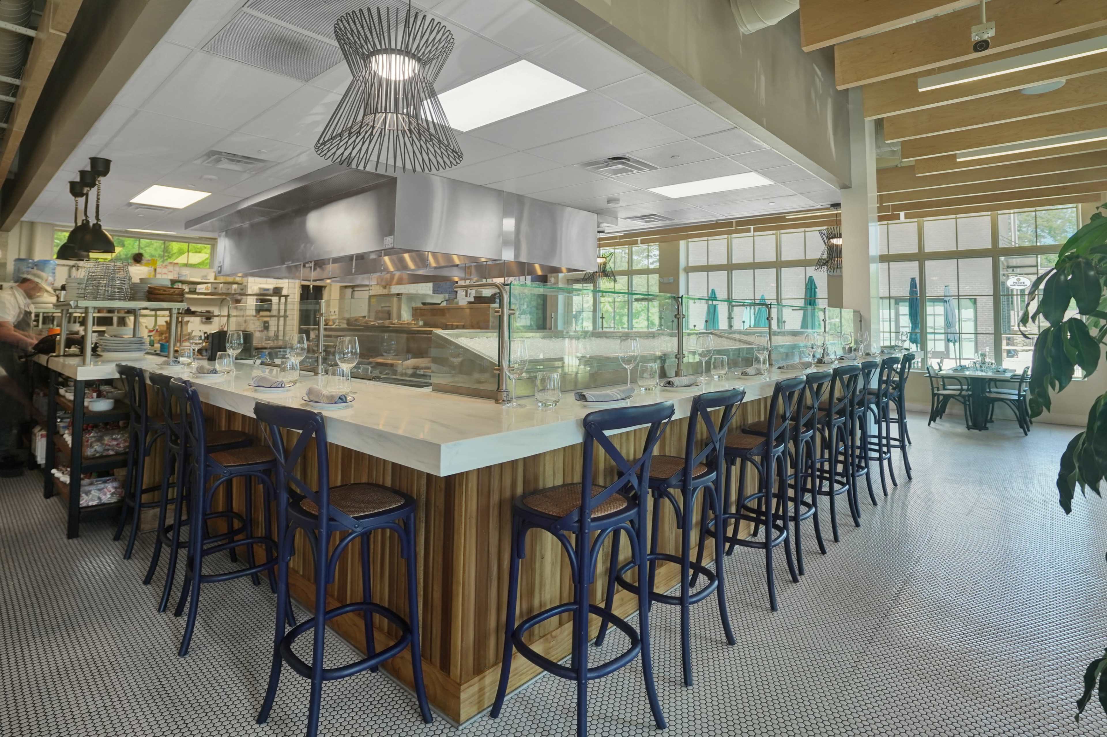 The image shows a modern restaurant interior with a large, central bar area featuring high stools and an open kitchen in the background.