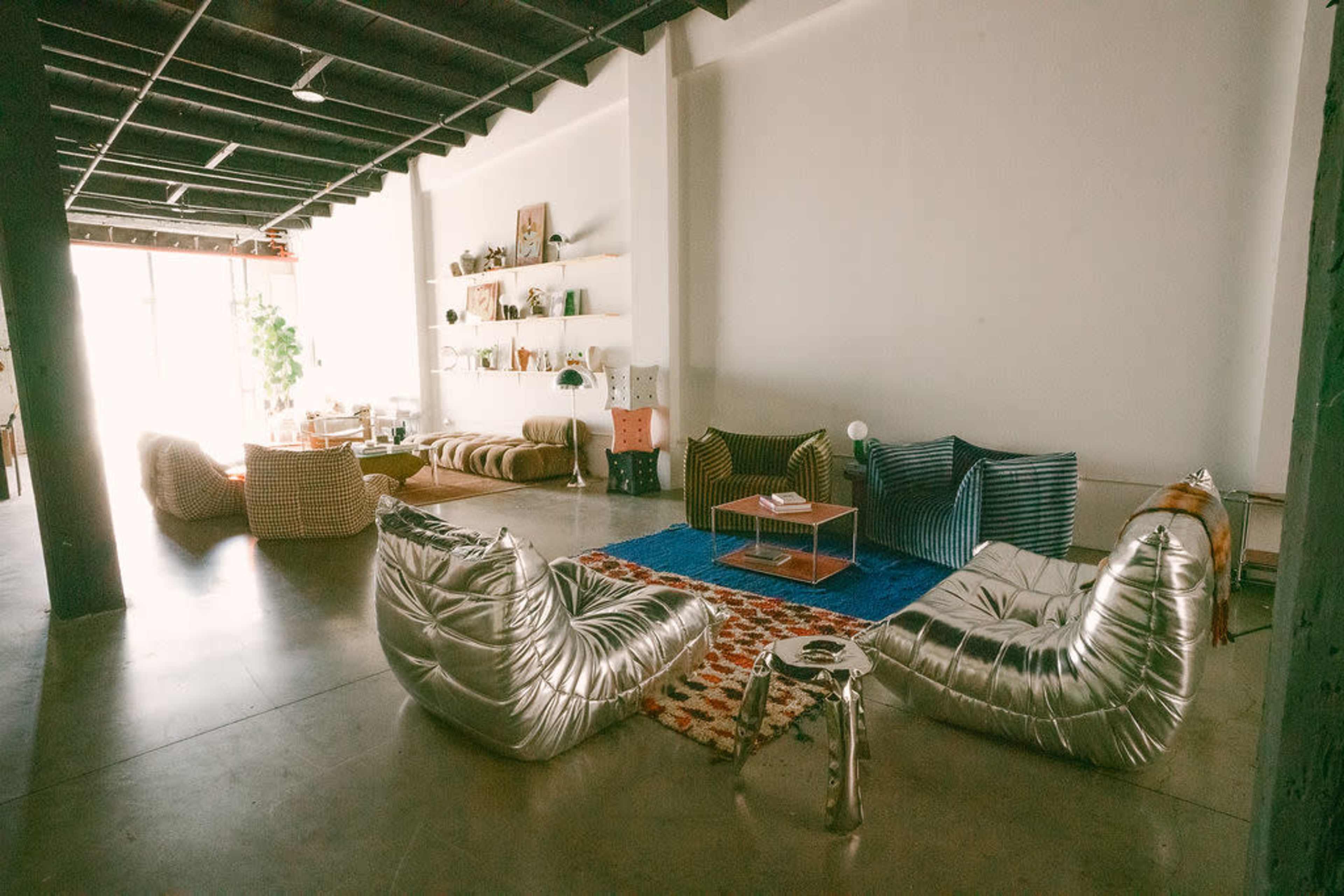 The image shows a spacious modern living area with several unique seating arrangements, a blue rug, and shelves displaying decor items.