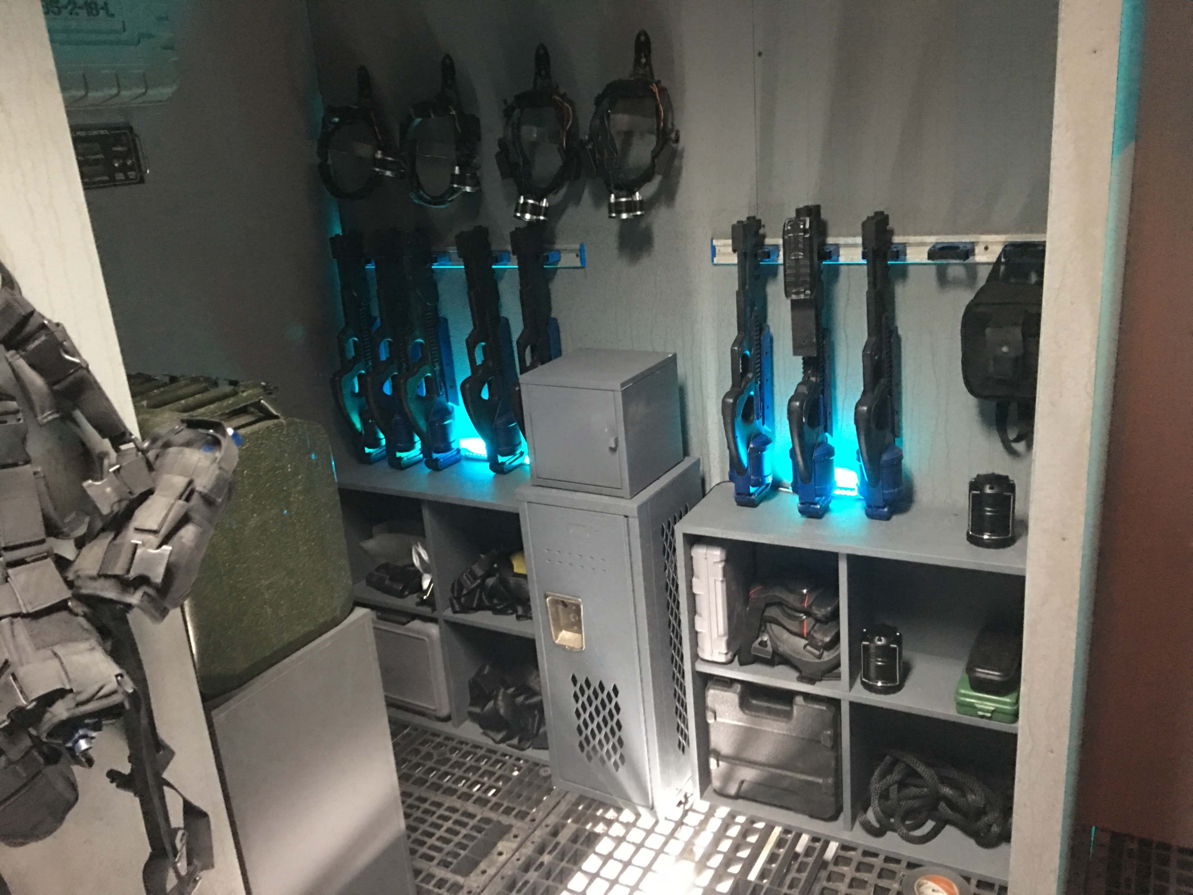 A storage area equipped with rifles, tactical gear, and a secure locker, all illuminated by blue lighting.