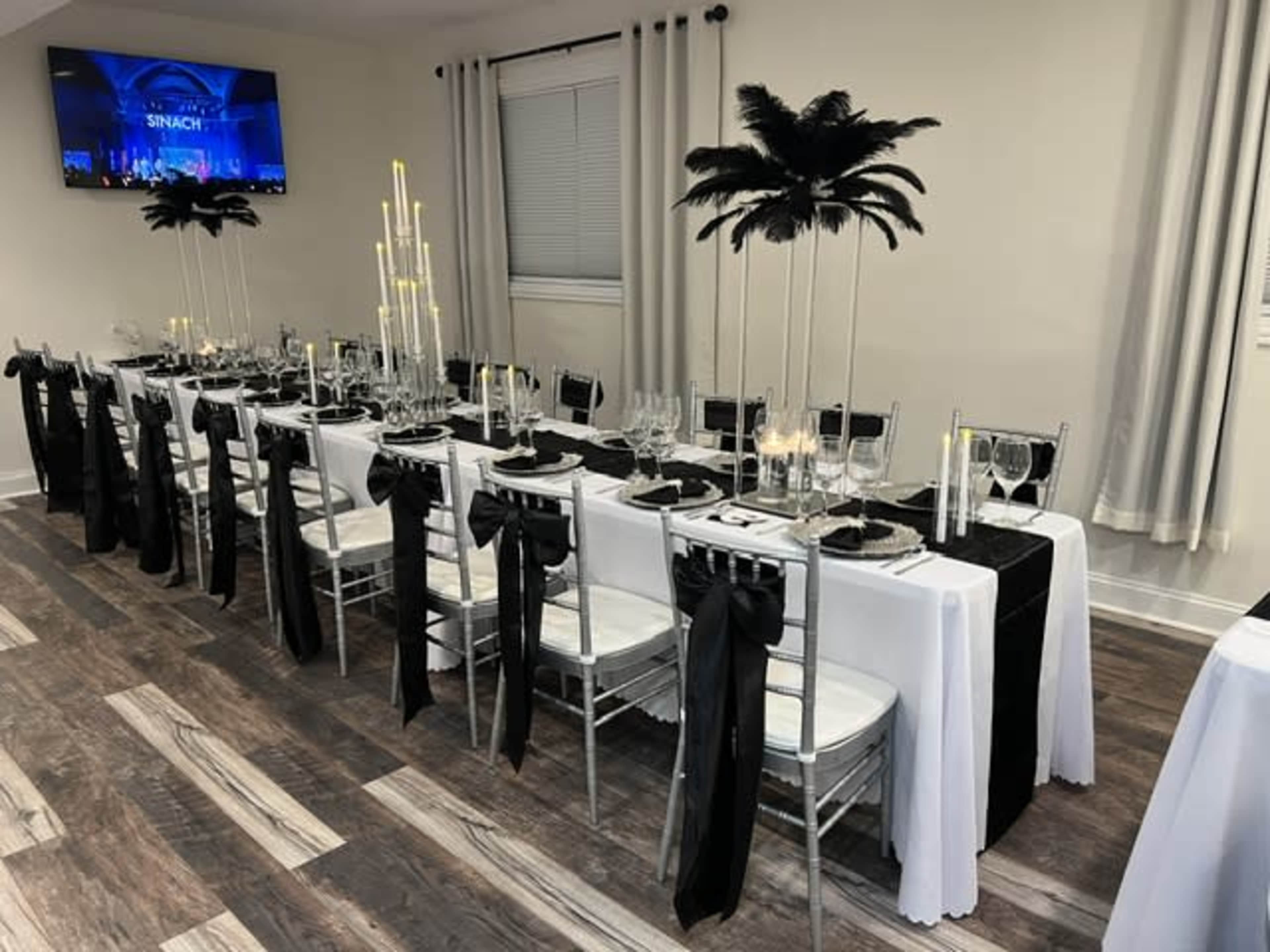 The image displays a banquet-style table set for a formal event, featuring black and white linens, tall centerpieces with feathers and candles, and elegant place settings.