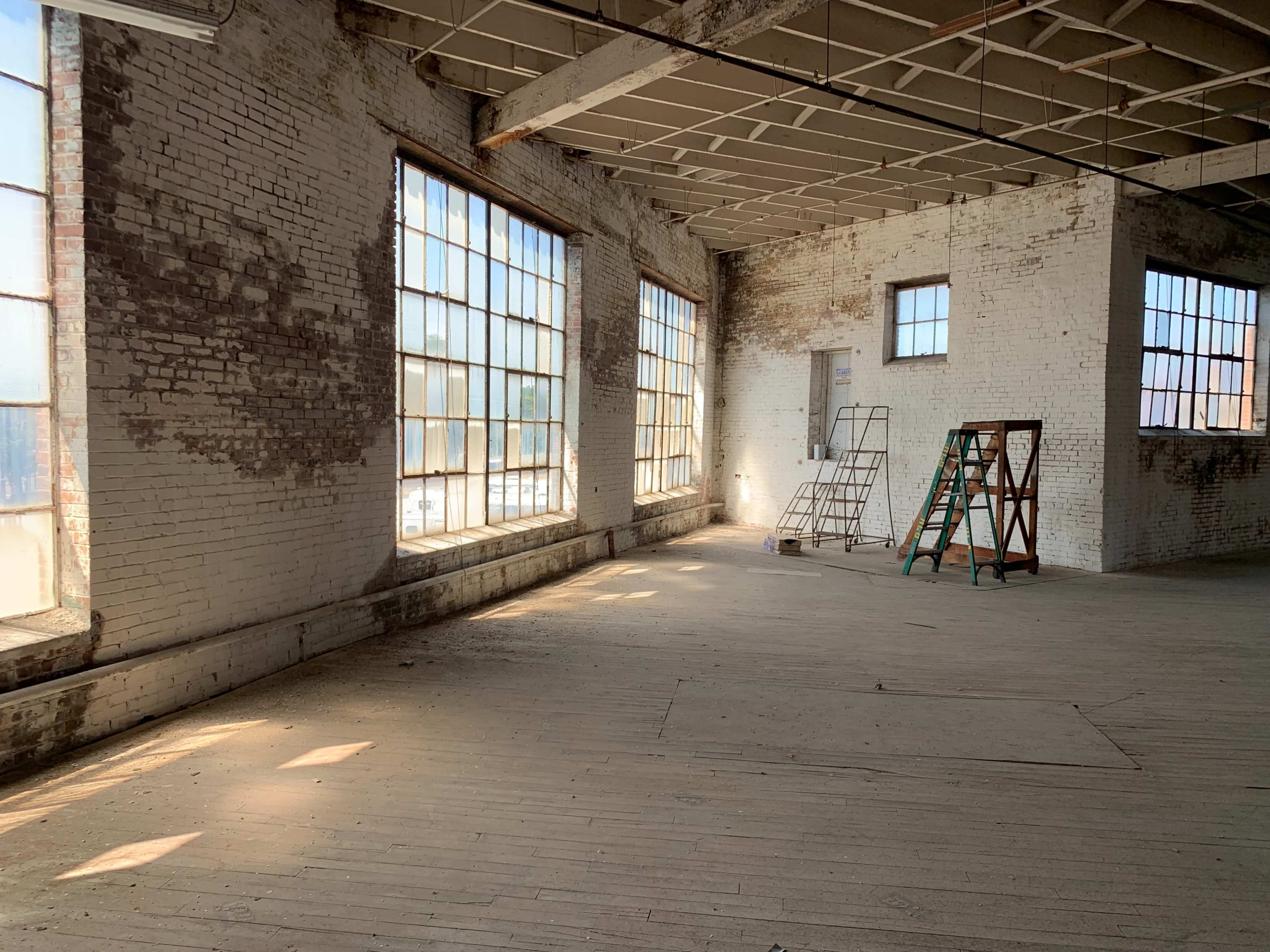 The image shows a spacious, vacant industrial room with large windows, exposed brick walls, and a ladder positioned near the center.