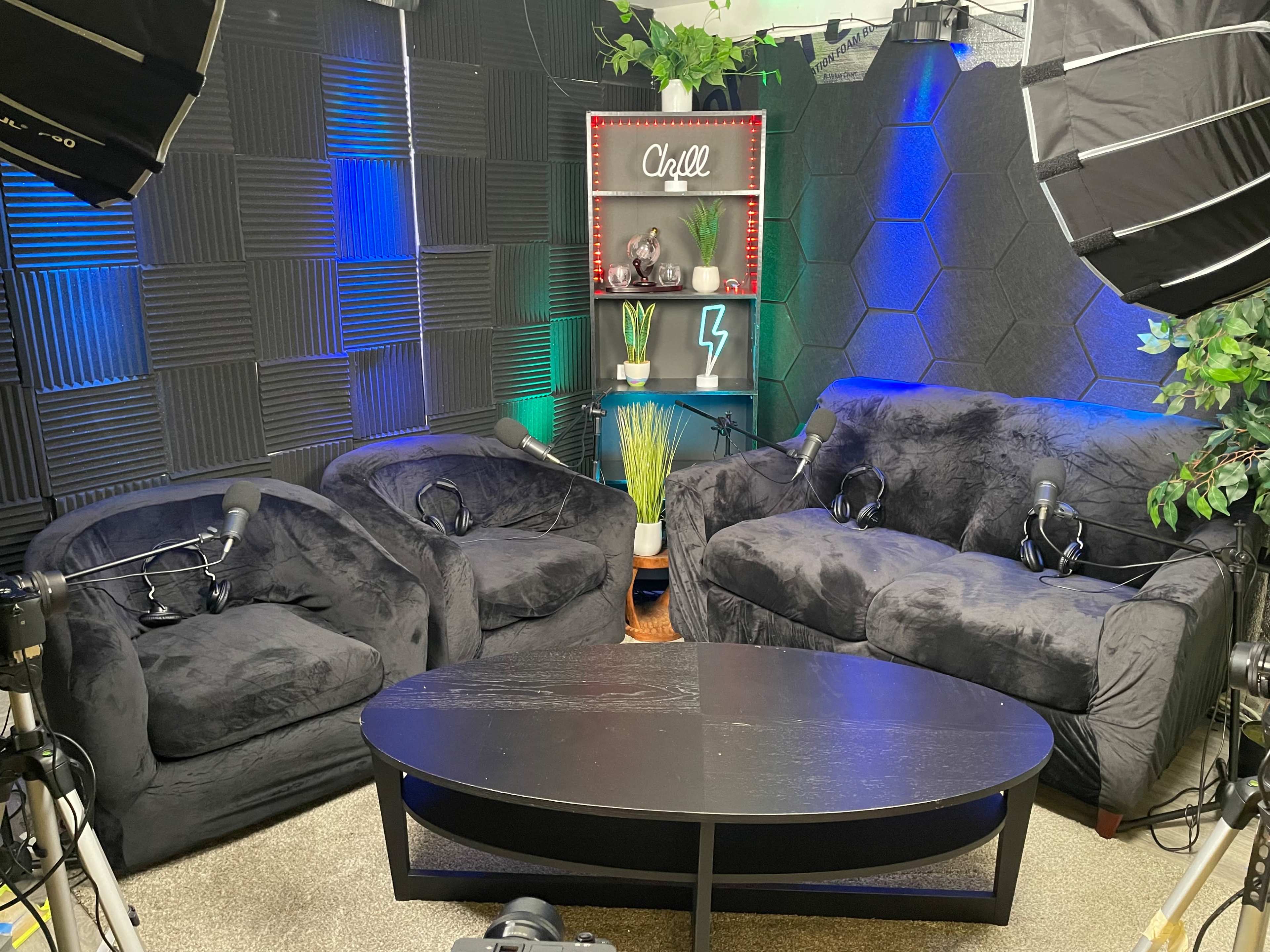 The image shows a cozy room setup with two black couches, a dark coffee table, and a backdrop featuring decorative plants and lighting, intended for a podcast or interview.