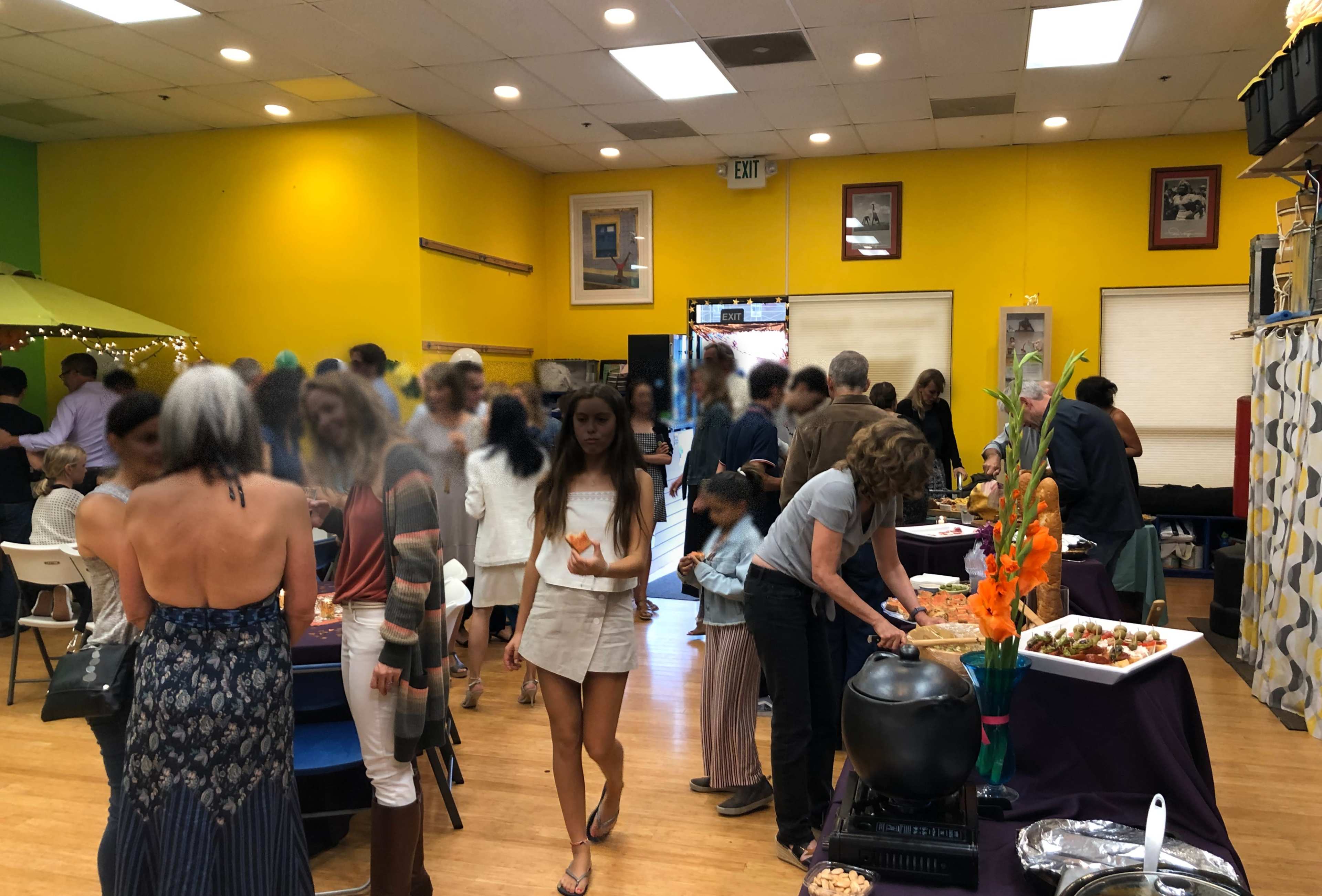 People gather in a brightly colored room filled with food tables and socializing activities during a community event.