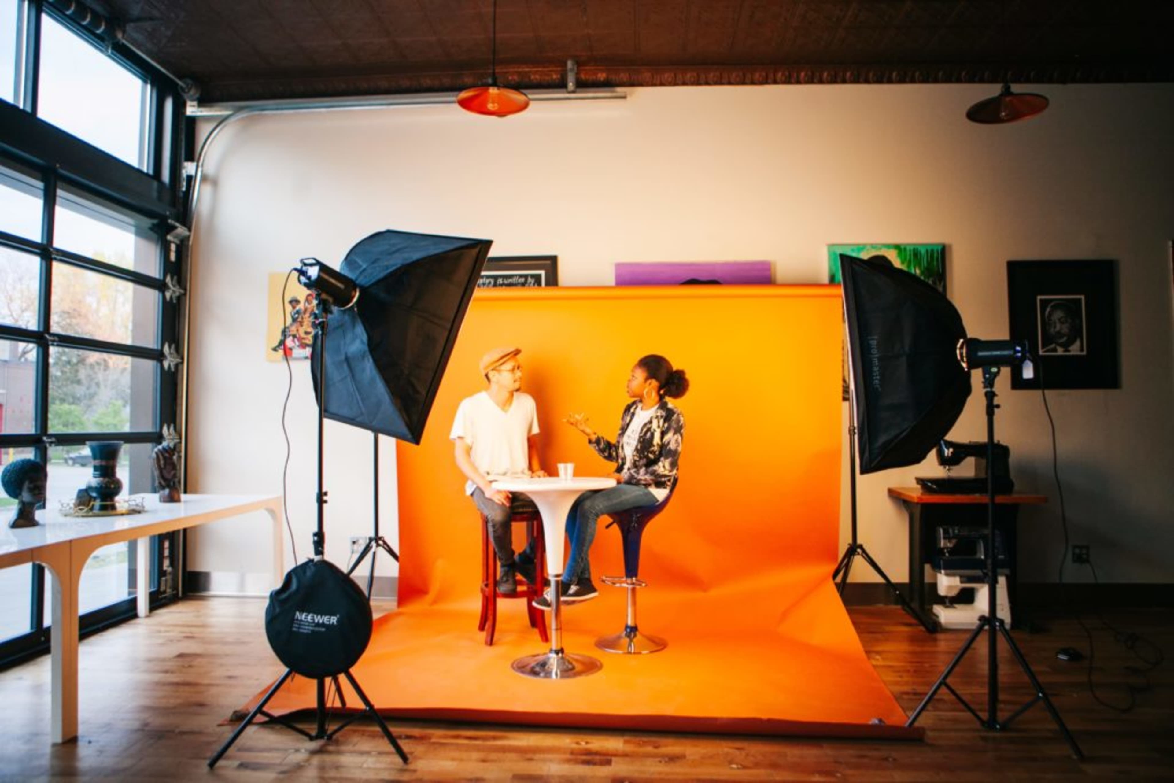 Two individuals are seated at a round table in front of an orange backdrop, surrounded by studio lighting equipment.
