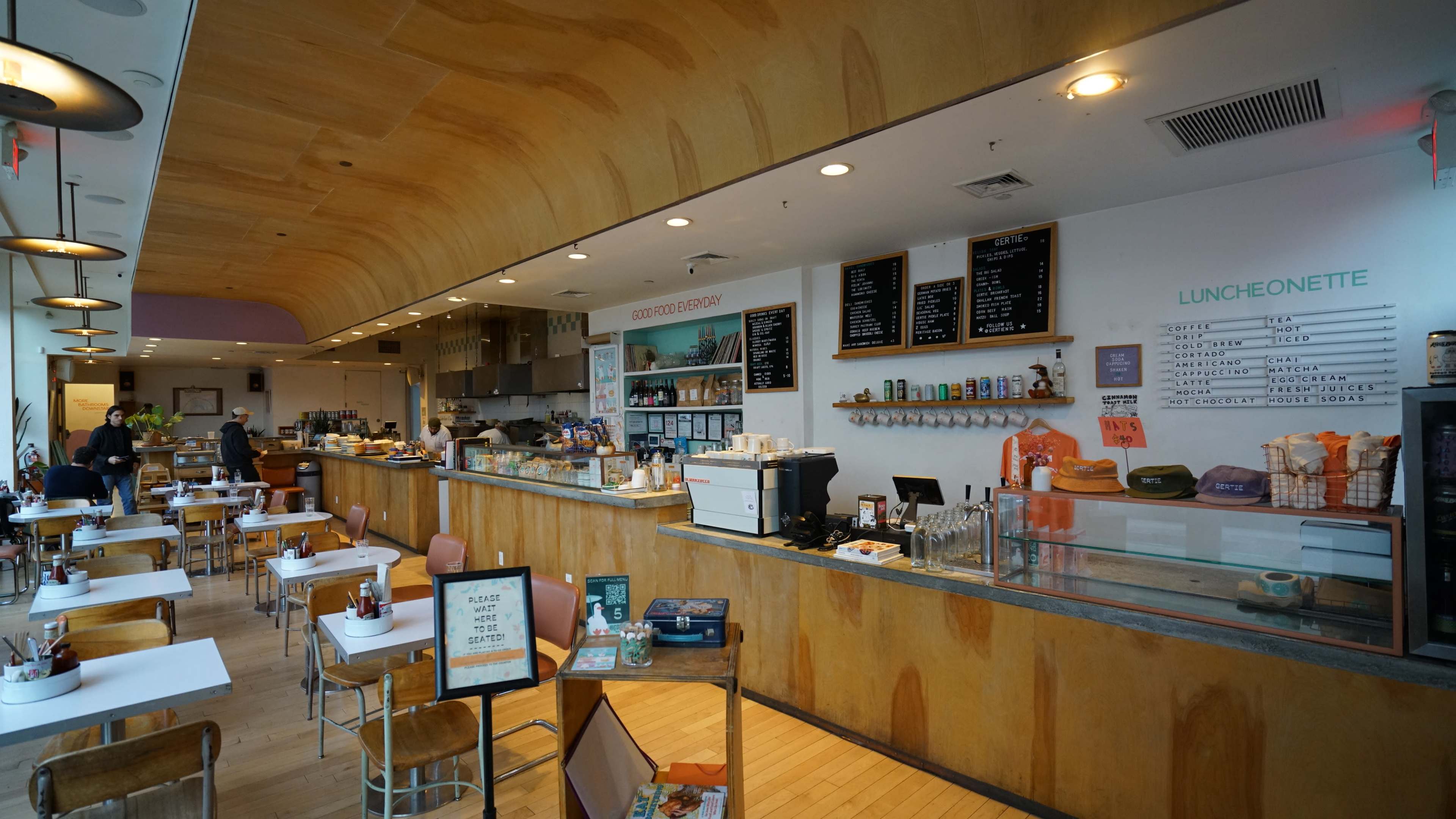 The image shows a modern café interior with wooden accents, a counter displaying food and drinks, and seating areas with tables and chairs.