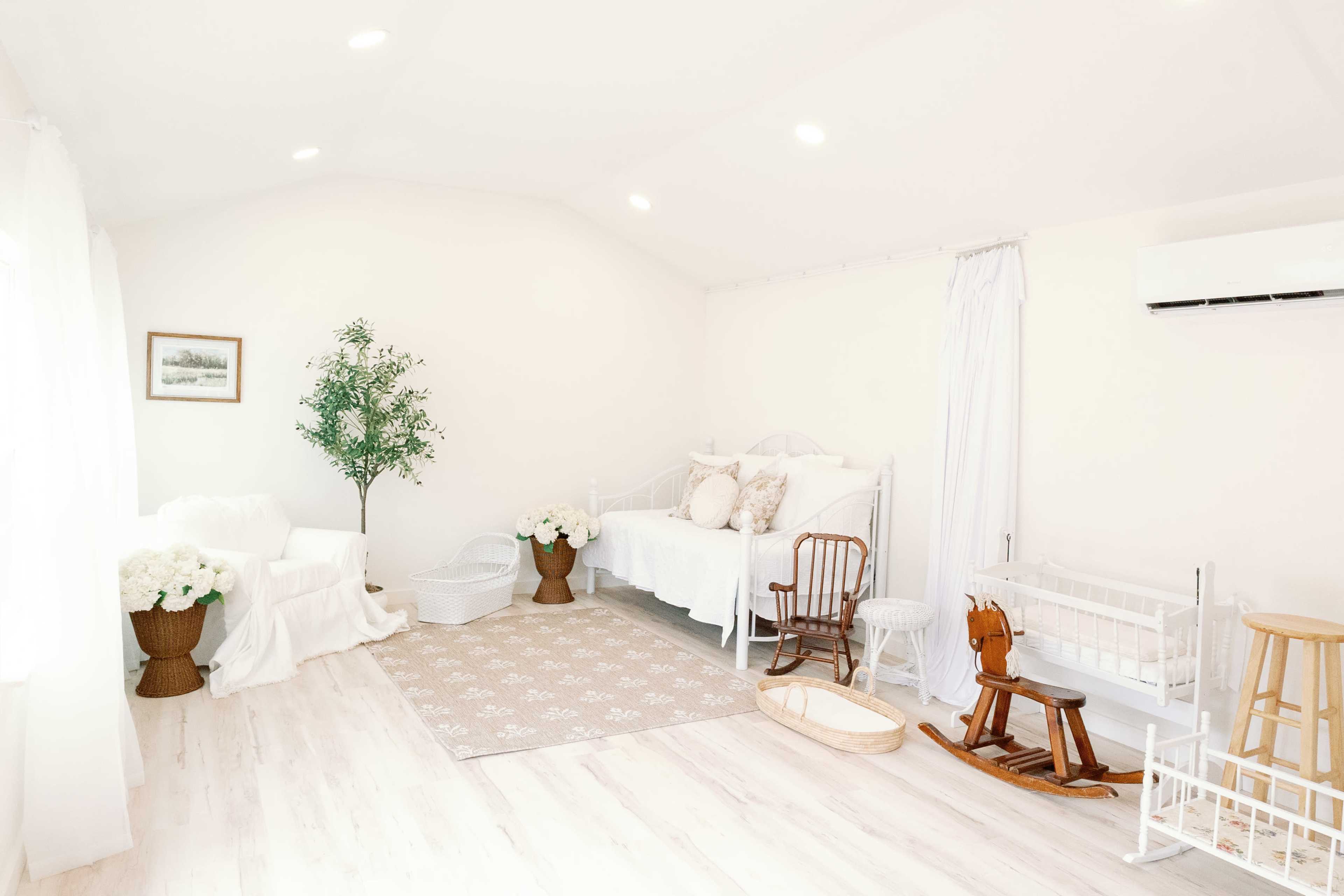 The image shows a bright, minimalist room featuring two beds, a rocking horse, and decorative plants.