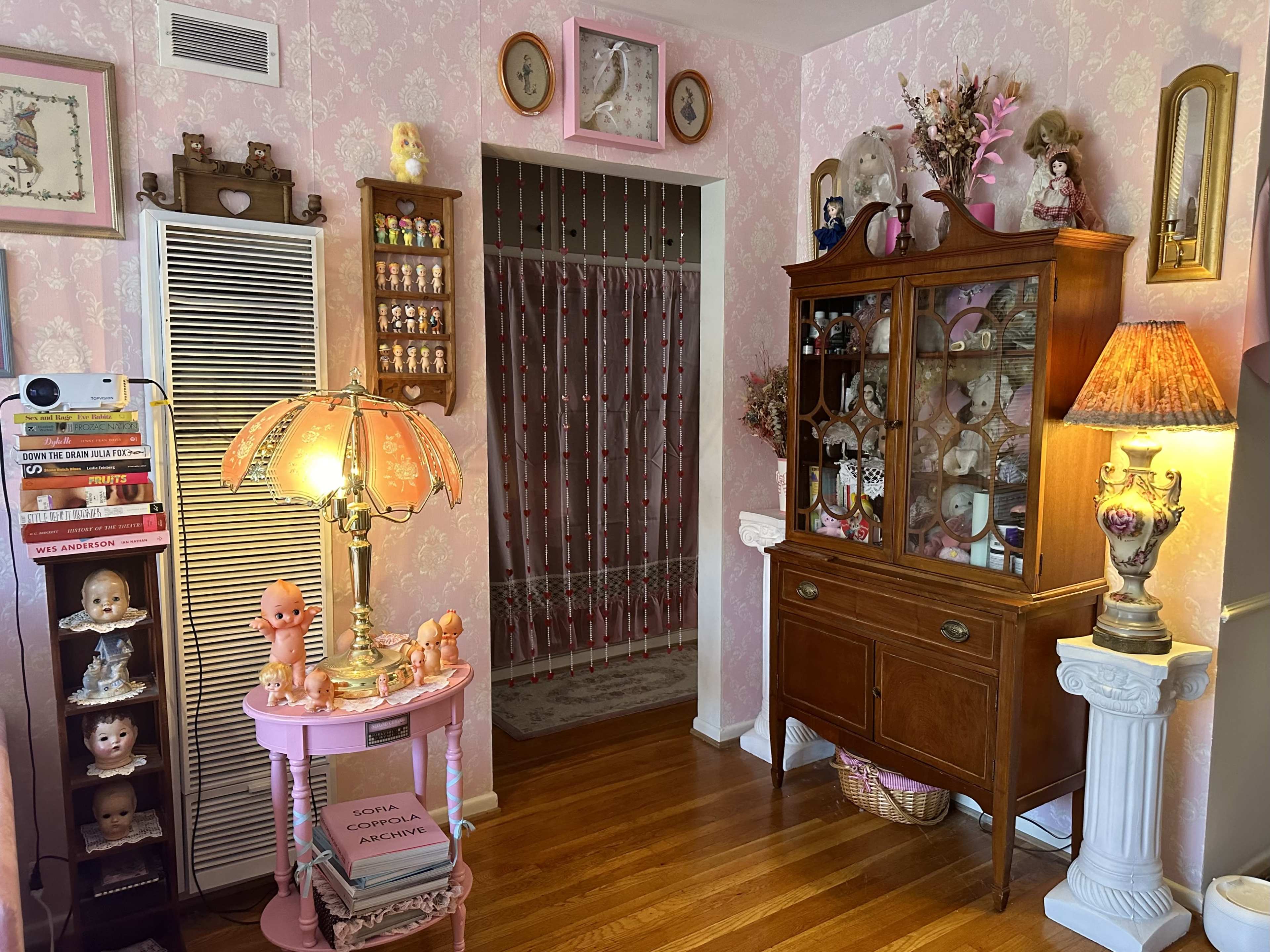The room features vintage decor with a pink wallpapered wall, a wooden cabinet displaying glassware, and several antique lamps and dolls arranged throughout.