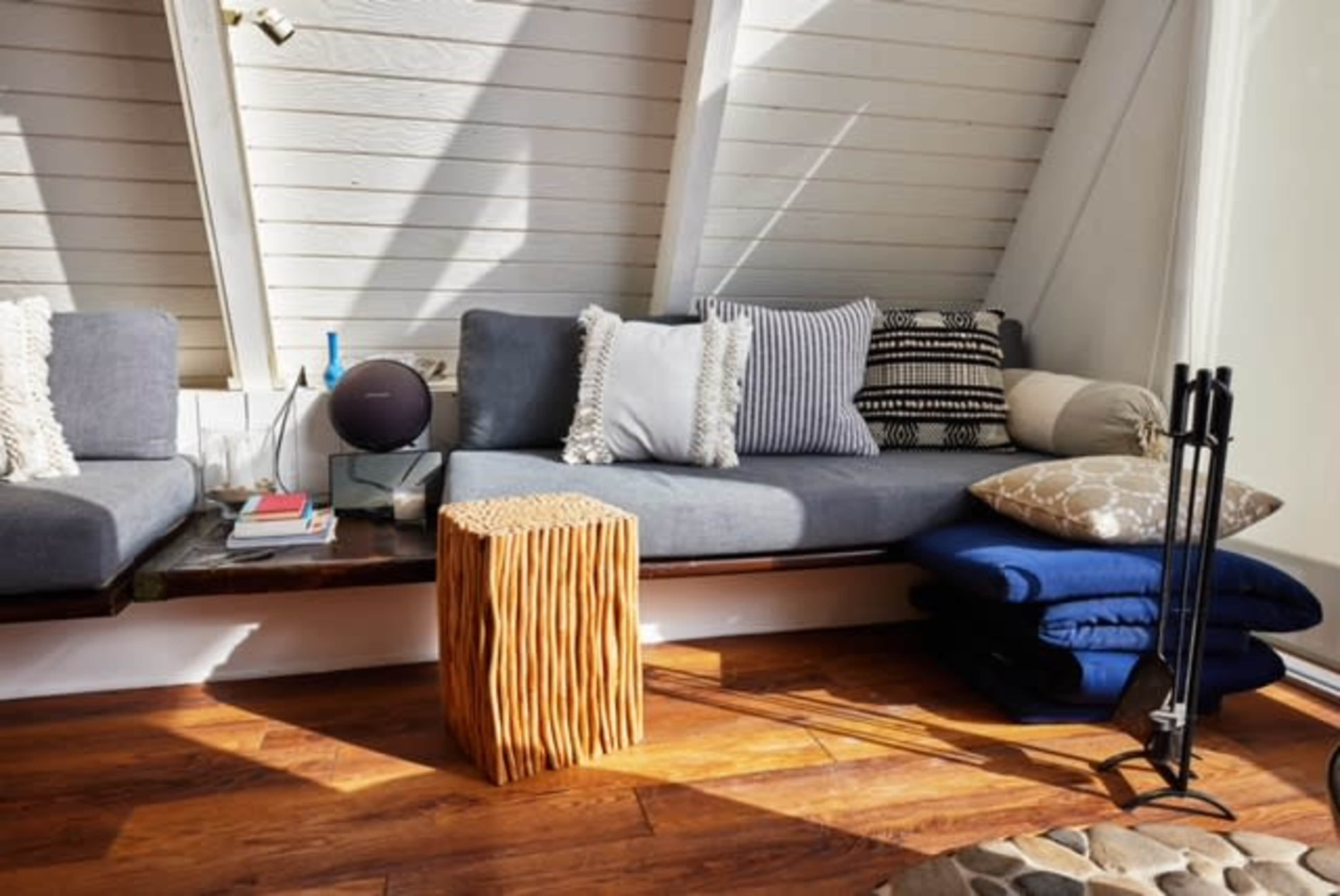 A cozy living space with a grey couch adorned with various pillows, a wooden side table, and stacked blue blankets on a polished wooden floor.