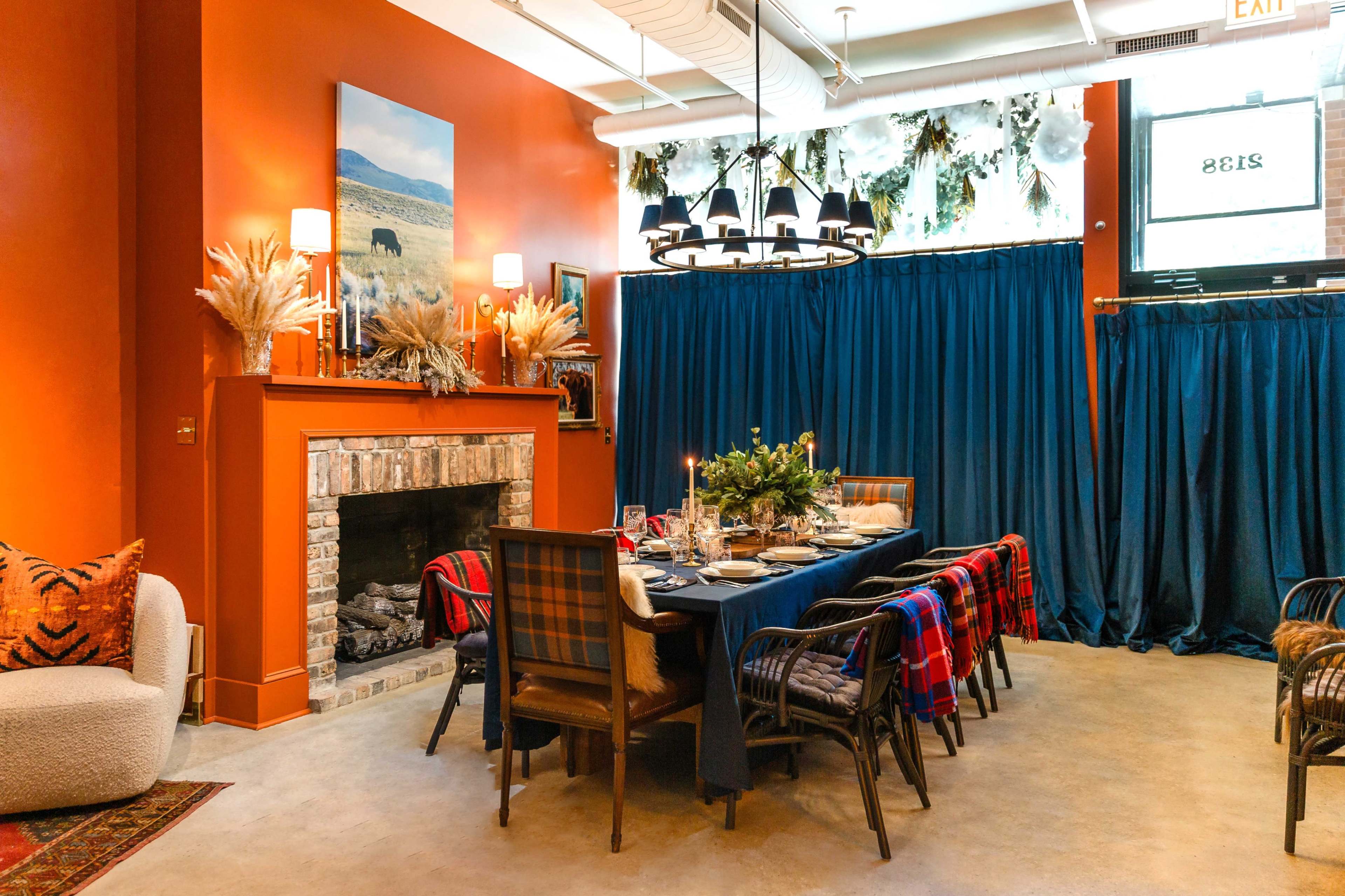 The image shows a cozy dining room with an orange wall, a large table set for a meal, and a fireplace adorned with decorative elements, all complemented by blue drapes and a contemporary light fixture.