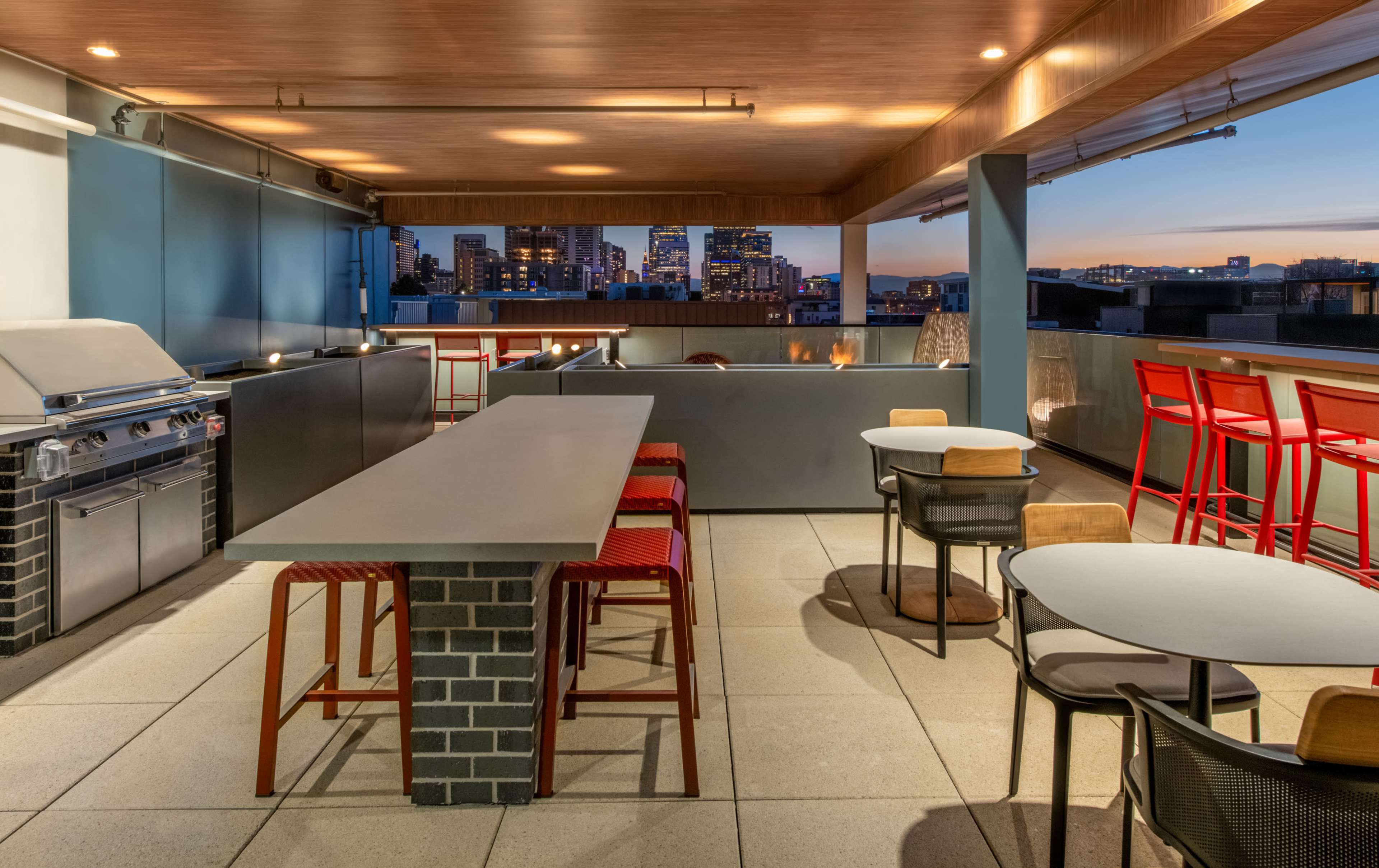 A modern rooftop terrace featuring a grill, bar seating, and tables with city skyline views at dusk.