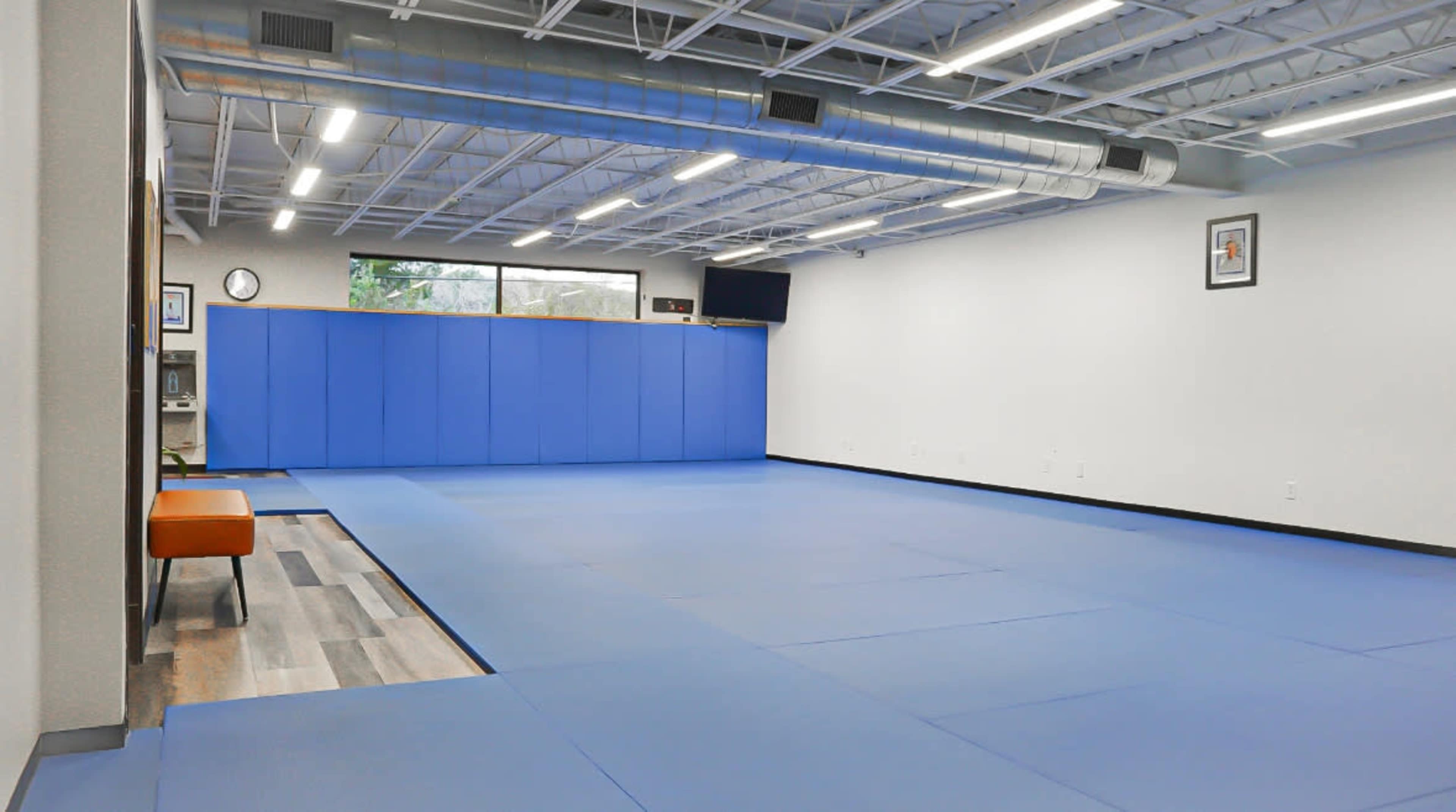 The image shows a large, empty training room with blue mats, a bench on the left, and a window revealing natural light.