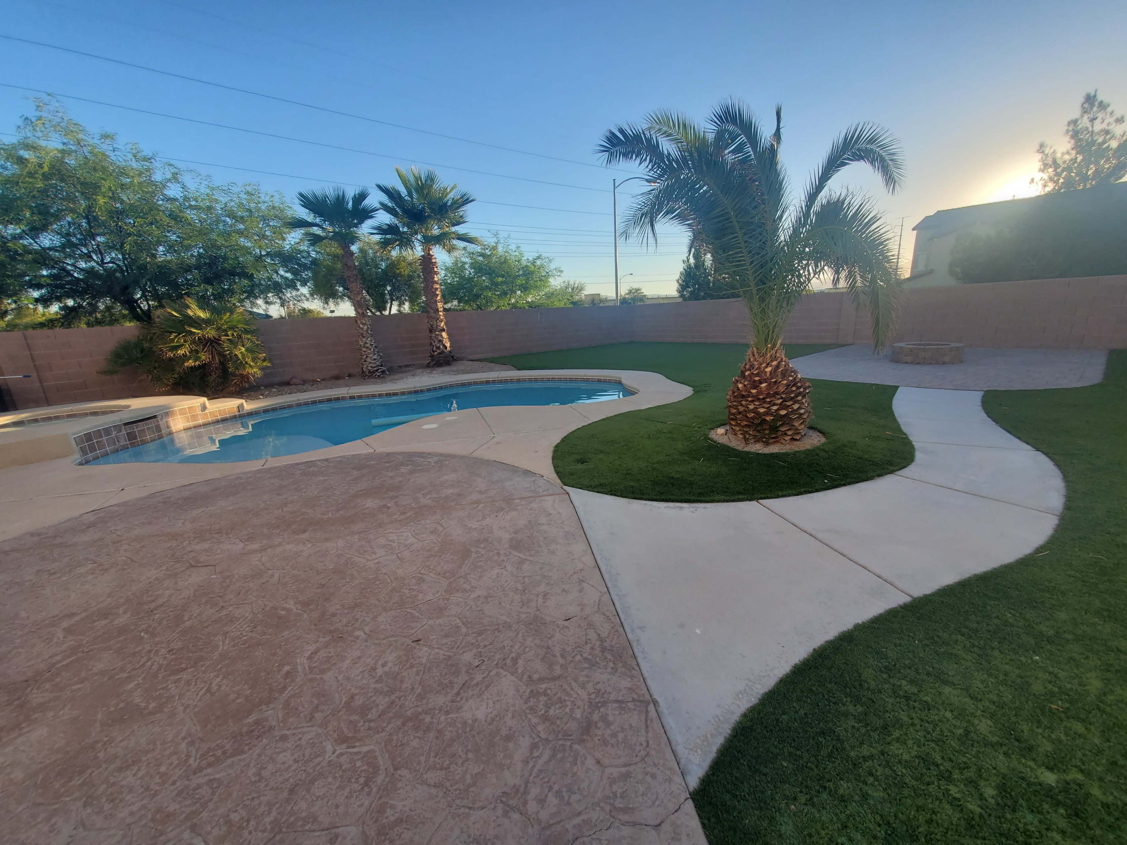 A backyard with a swimming pool surrounded by landscaped grass, palm trees, and a stone fire pit.