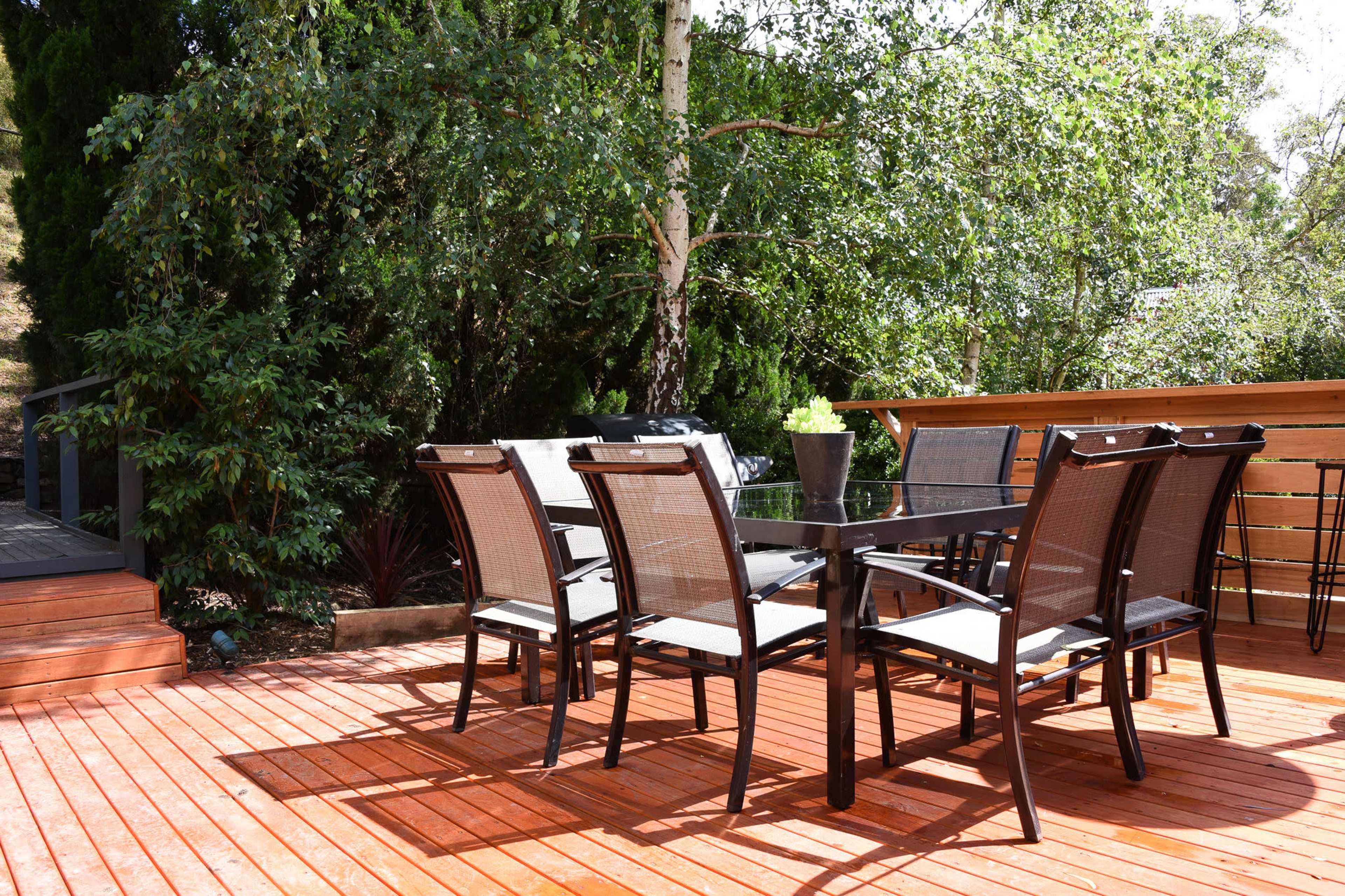 A wooden deck features a table surrounded by six black mesh chairs, set in a lush green garden.