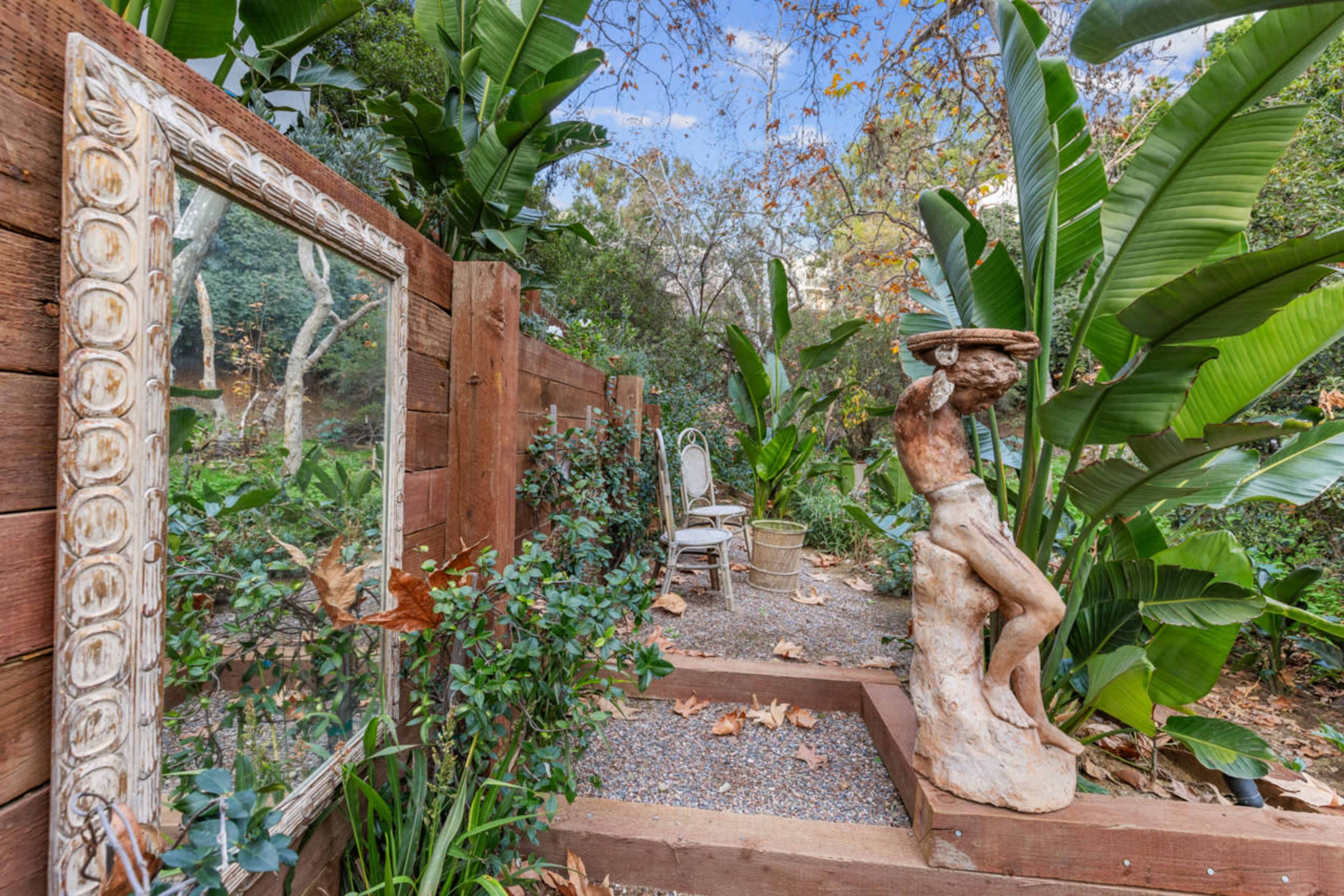 A garden scene featuring a wooden fence, a mirror framed in ornate style, a stone statue of a figure holding a dish, and a chair surrounded by greenery and plants.
