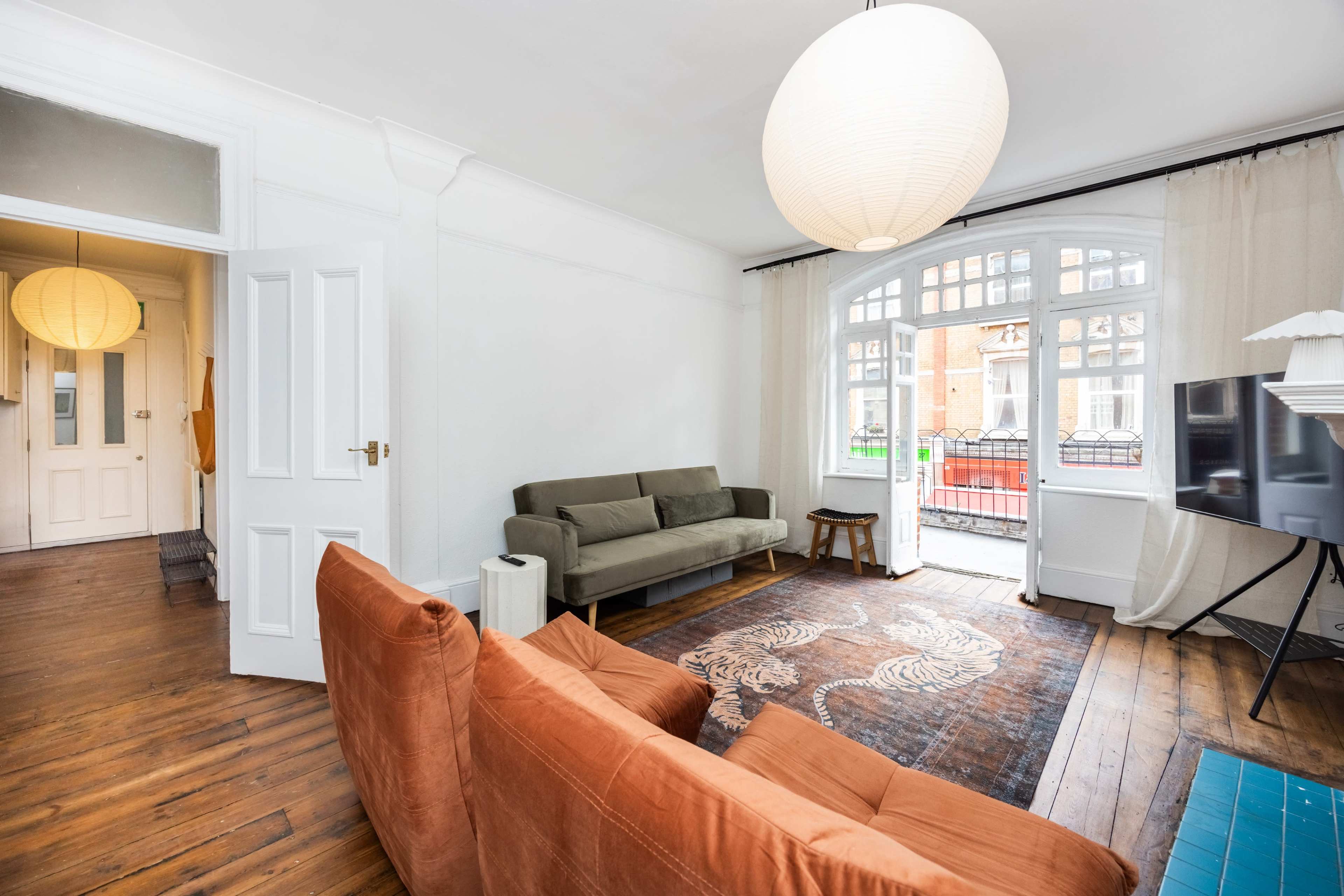A living room features a green sofa, an orange couch, a round paper lantern hanging from the ceiling, and a television on the wall, with large windows providing natural light.