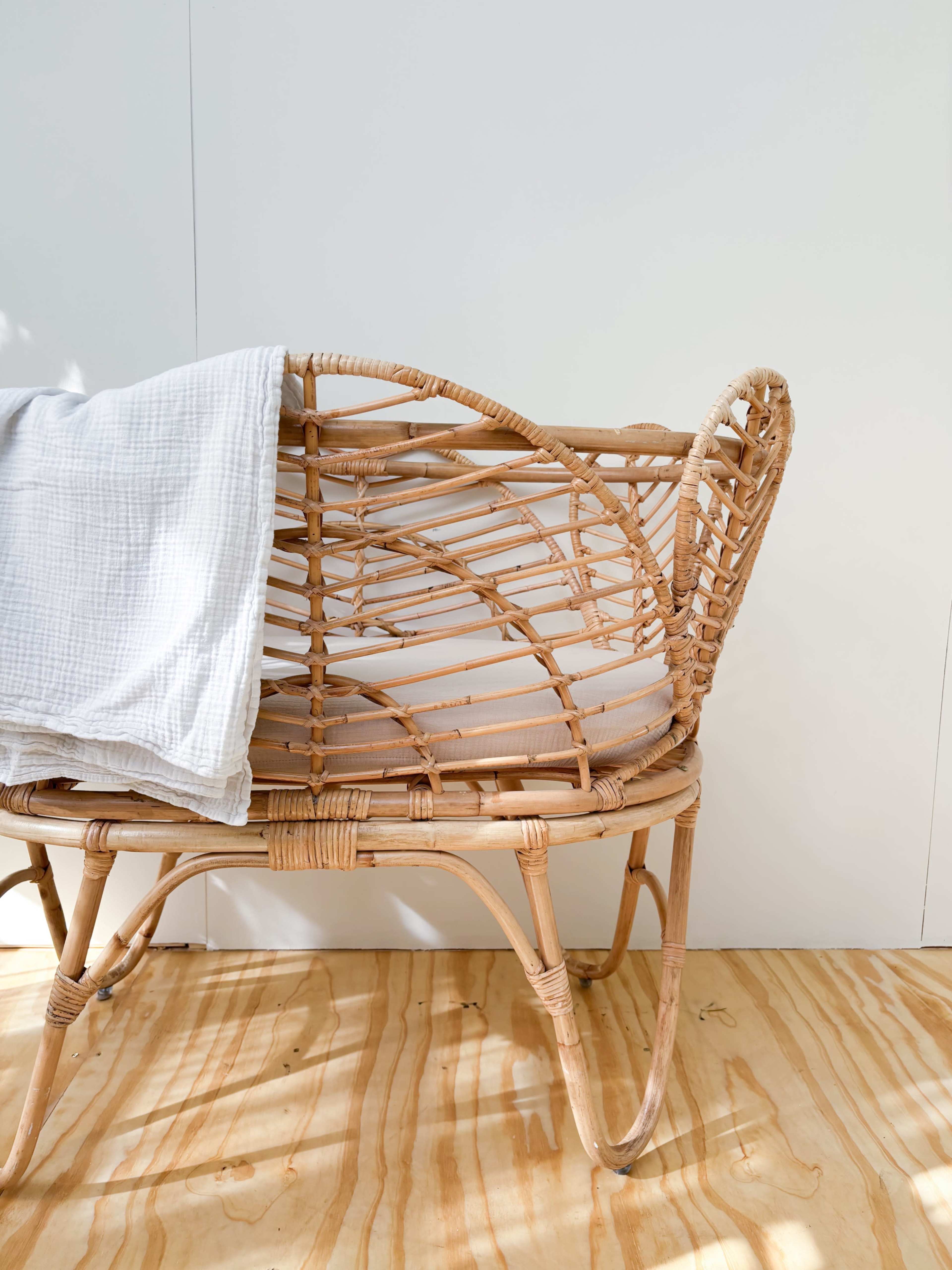 A woven rattan chair with a white blanket draped over it is positioned against a plain white wall.