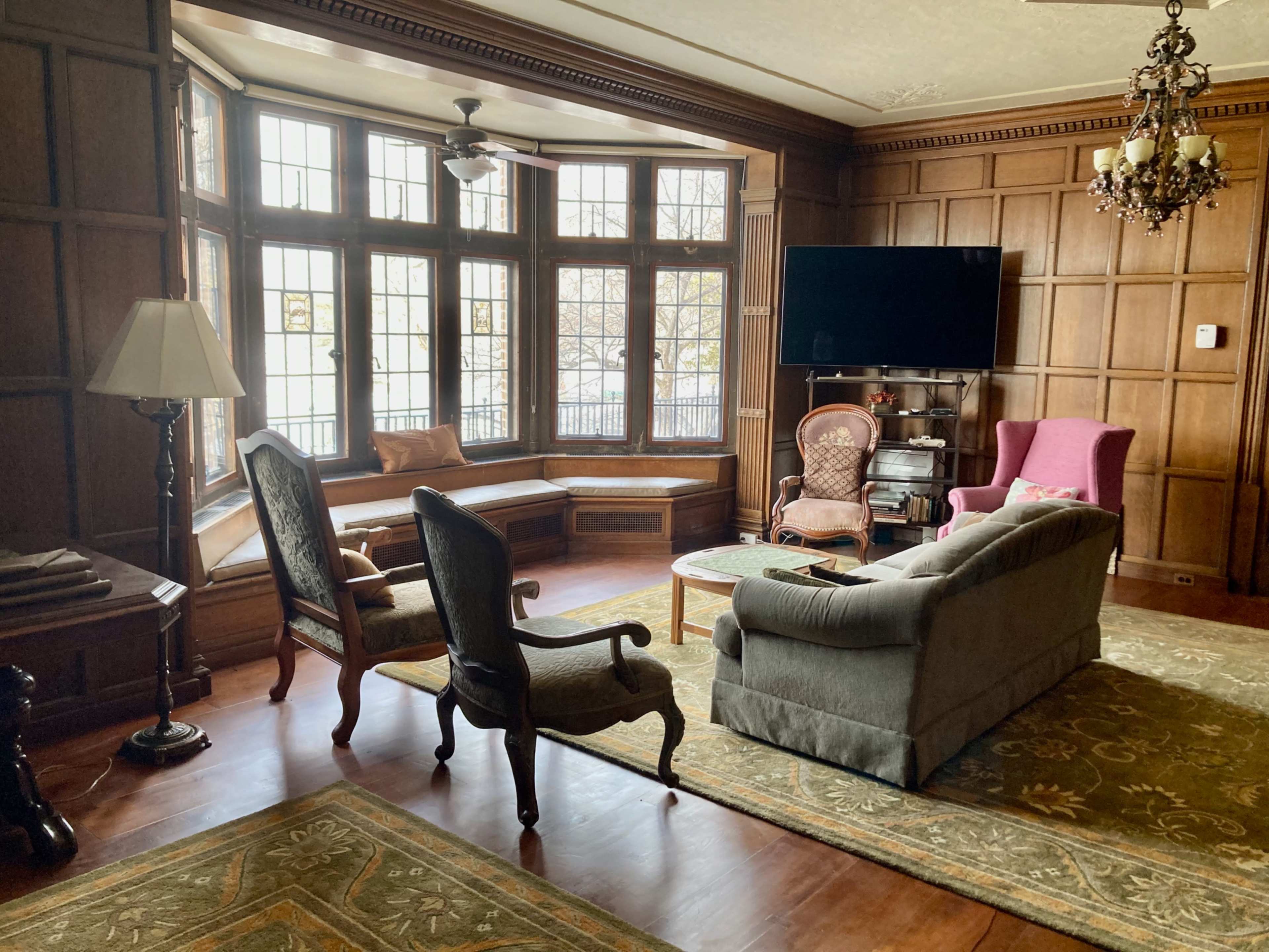 The living room features wooden paneling, large windows, and a combination of vintage furniture, including two armchairs and a sofa.