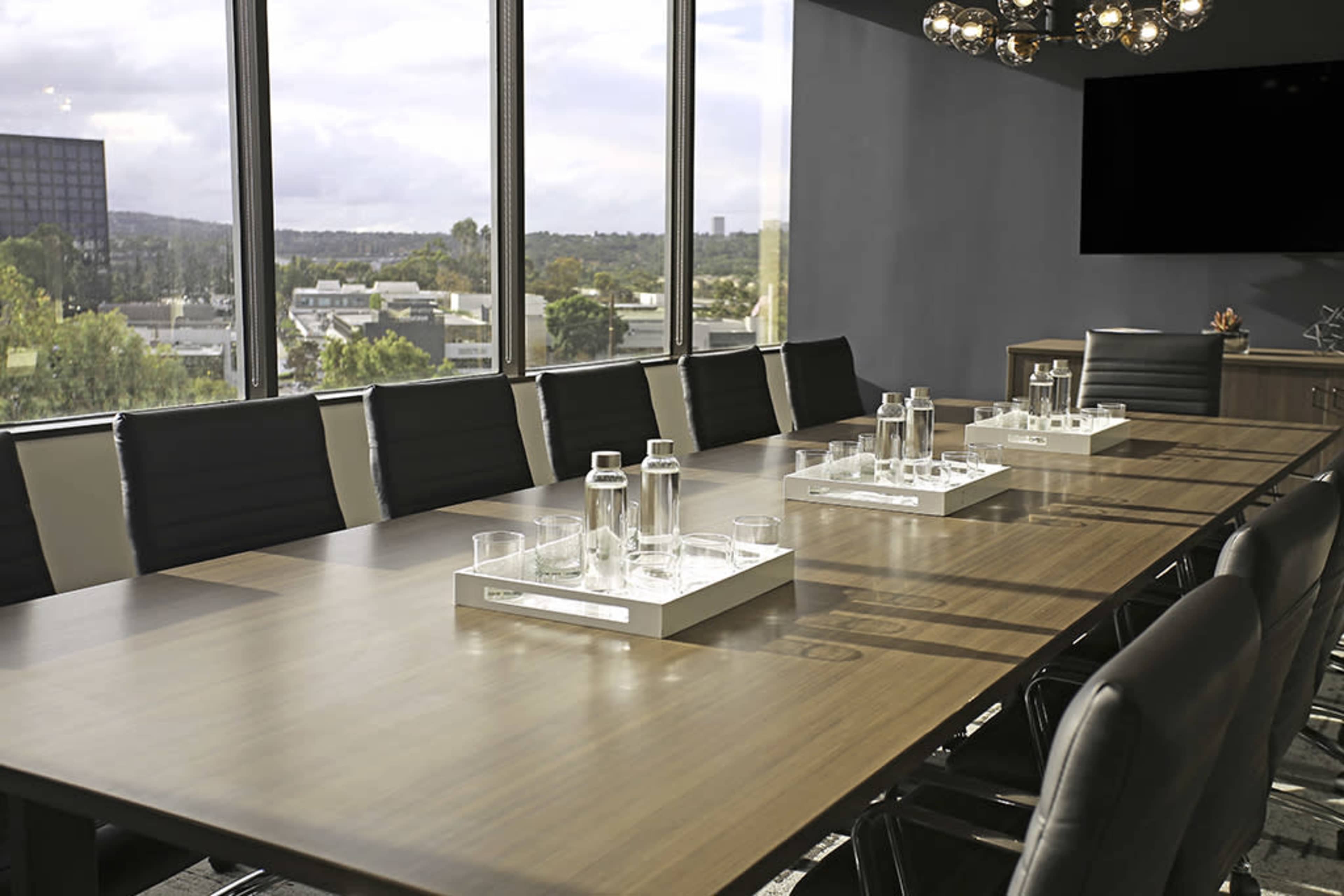 A long, modern conference table is set with glass water carafes and cups, surrounded by black and white chairs, with large windows providing a view of the cityscape.