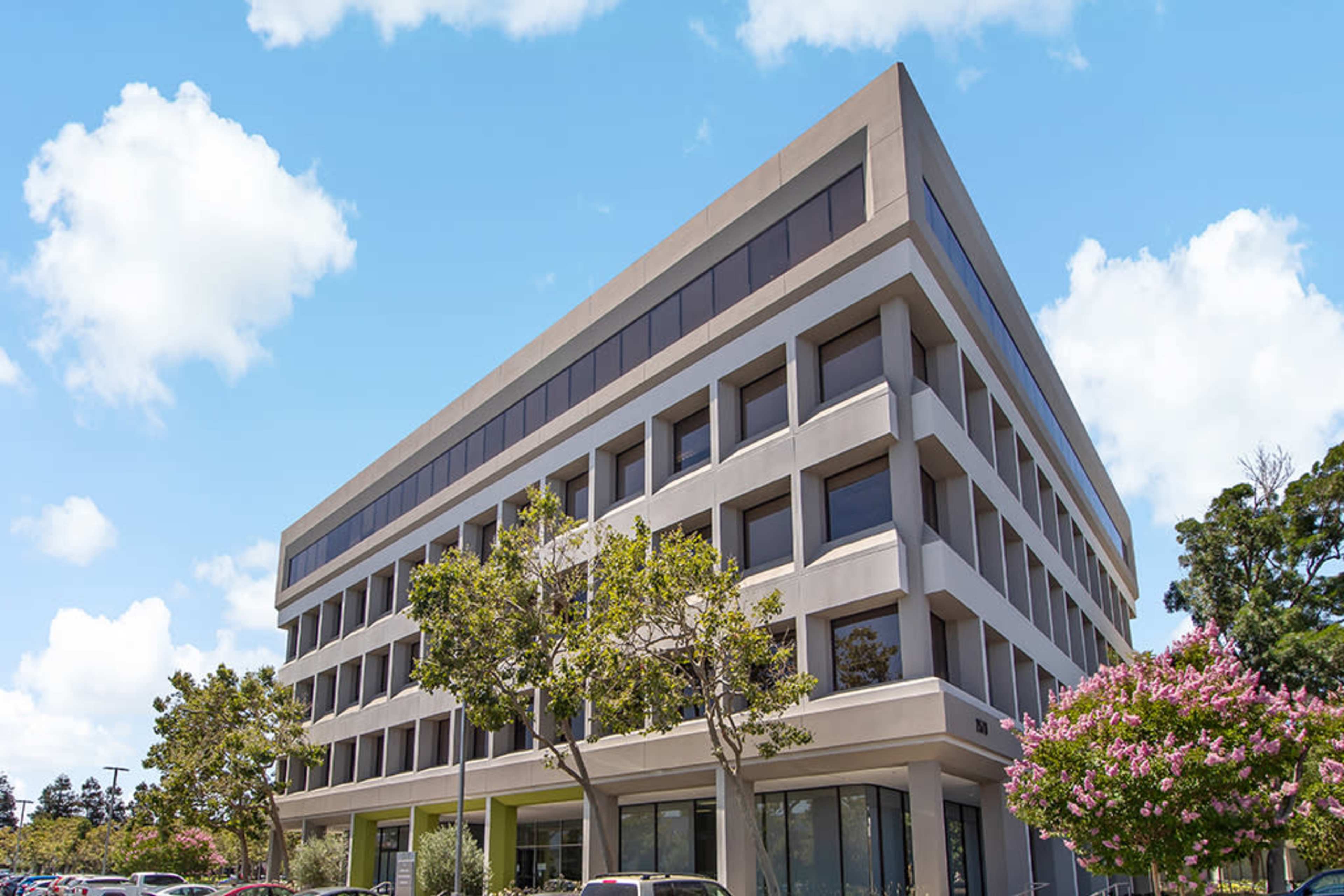 A modern five-story building with a flat roof and large windows is surrounded by trees and parked cars.