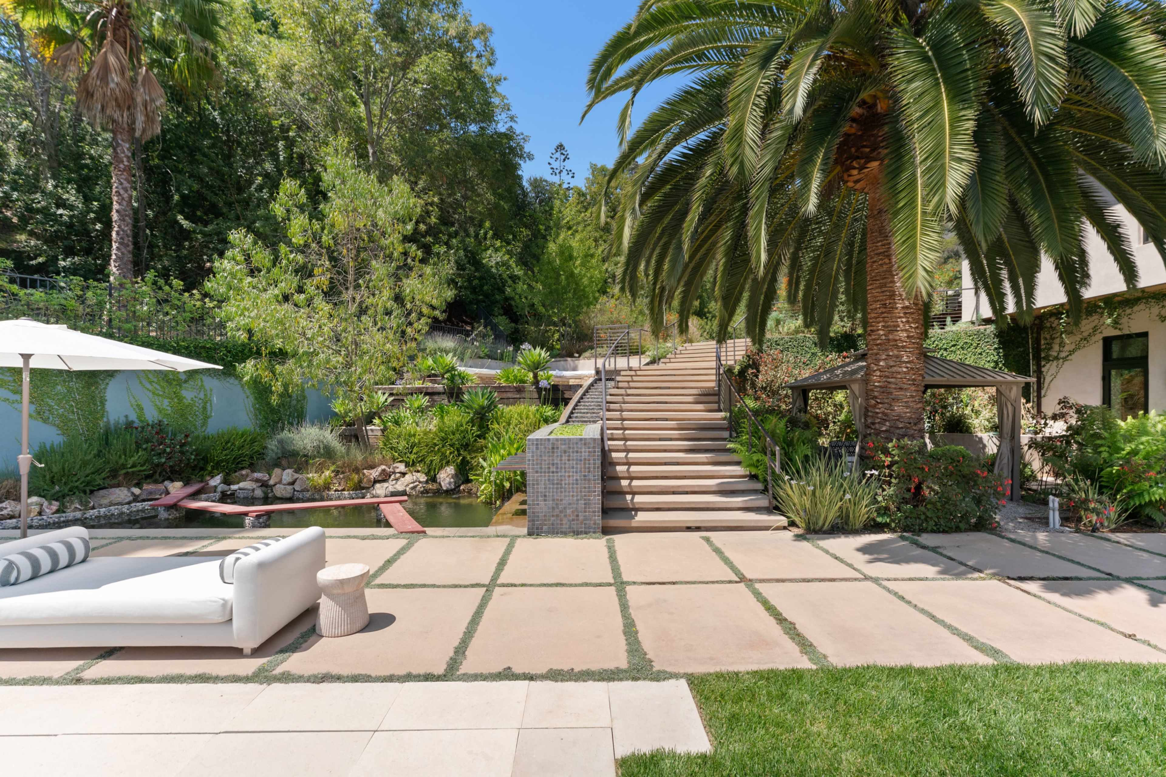 The image shows a landscaped outdoor area featuring a patio with a white sofa, stone pathways, staircases leading to lush greenery, and tall palm trees.