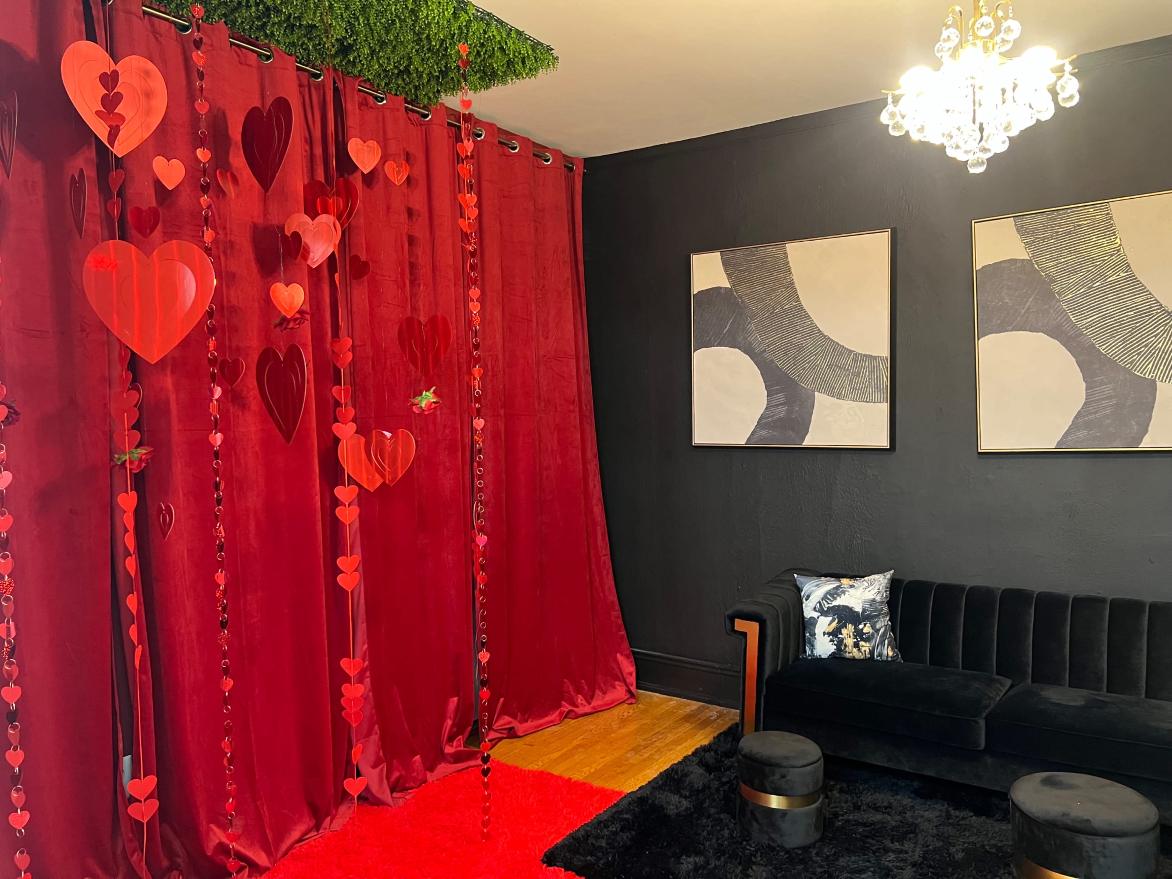 The image shows a cozy interior with a red curtain decorated with hanging hearts, a black sofa, and modern artwork on the walls.