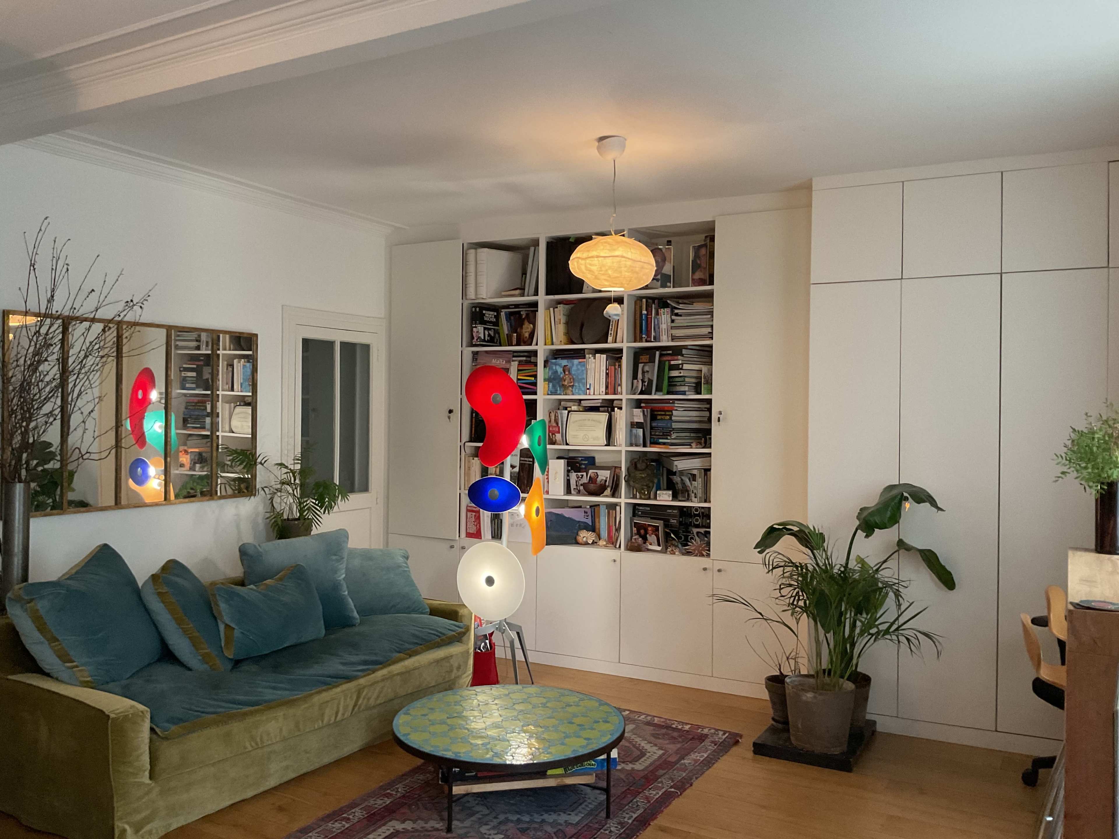 The image shows a cozy living room with a green sofa, a decorative round table, bookshelves filled with items, and several indoor plants.