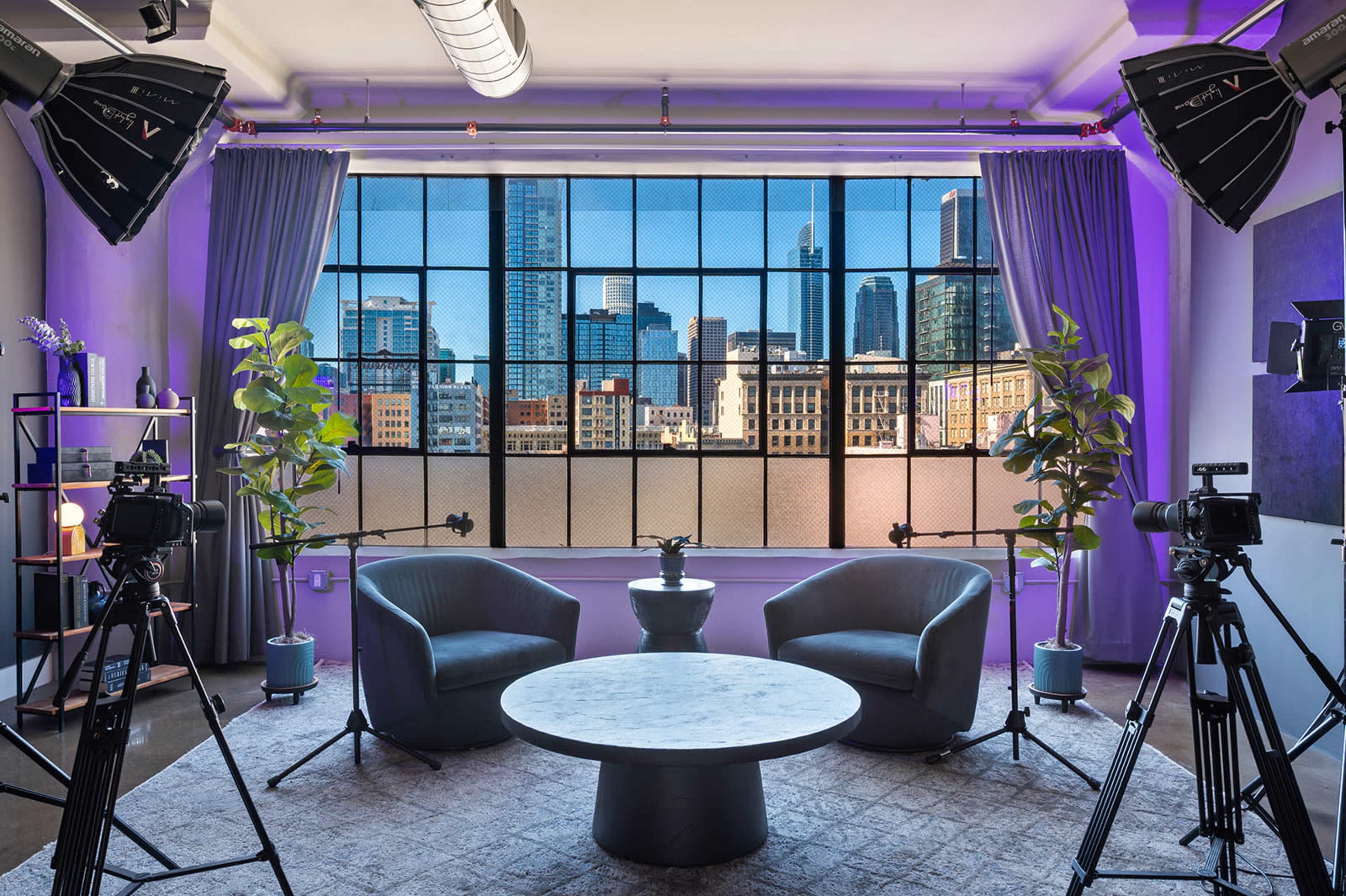 The image shows a modern filming setup featuring two chairs and a circular table, framed by large windows with a city skyline in the background.