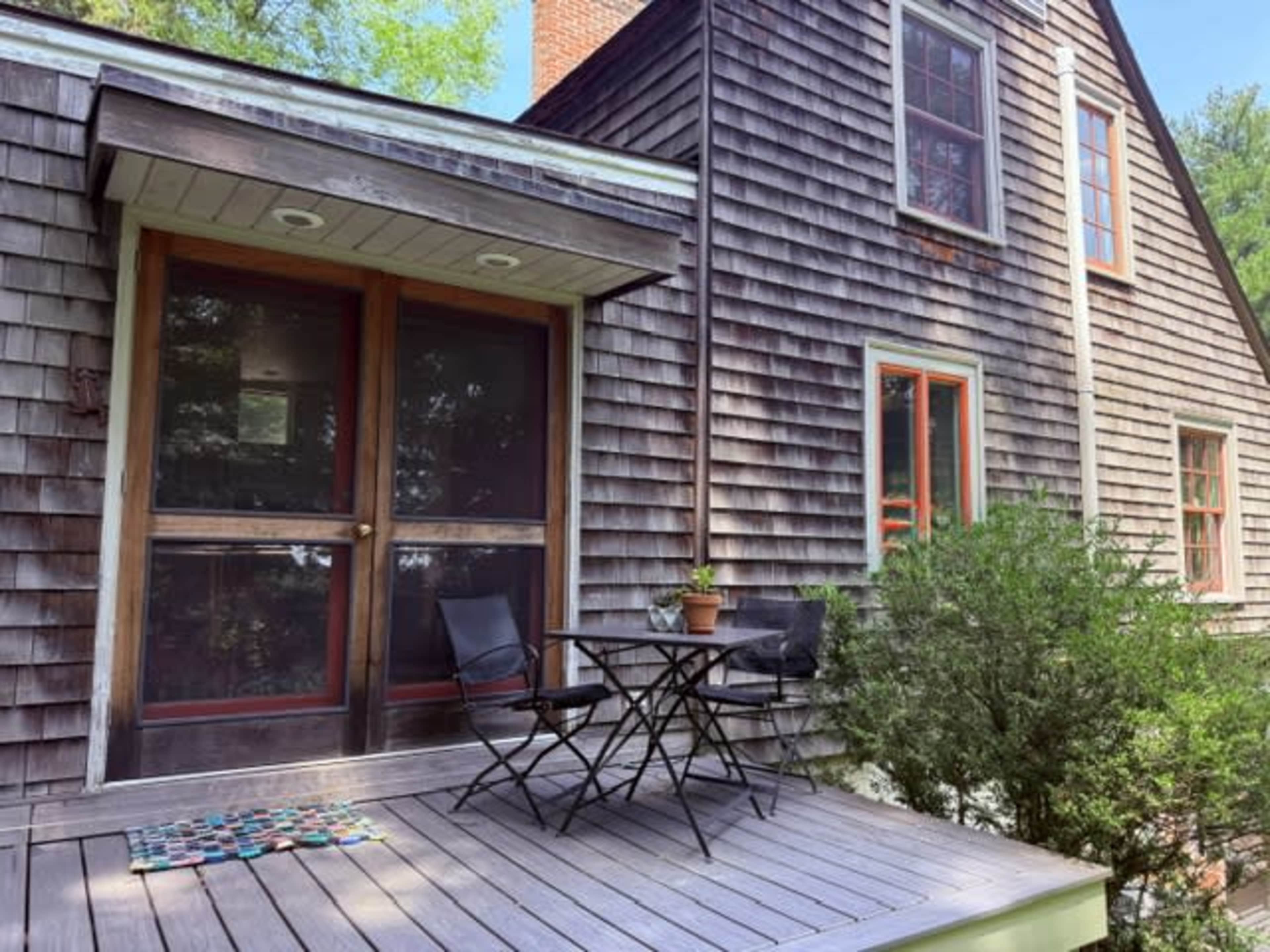 The image shows a wooden deck with a small table and two chairs, adjacent to a house with shingled siding and red-trimmed windows.