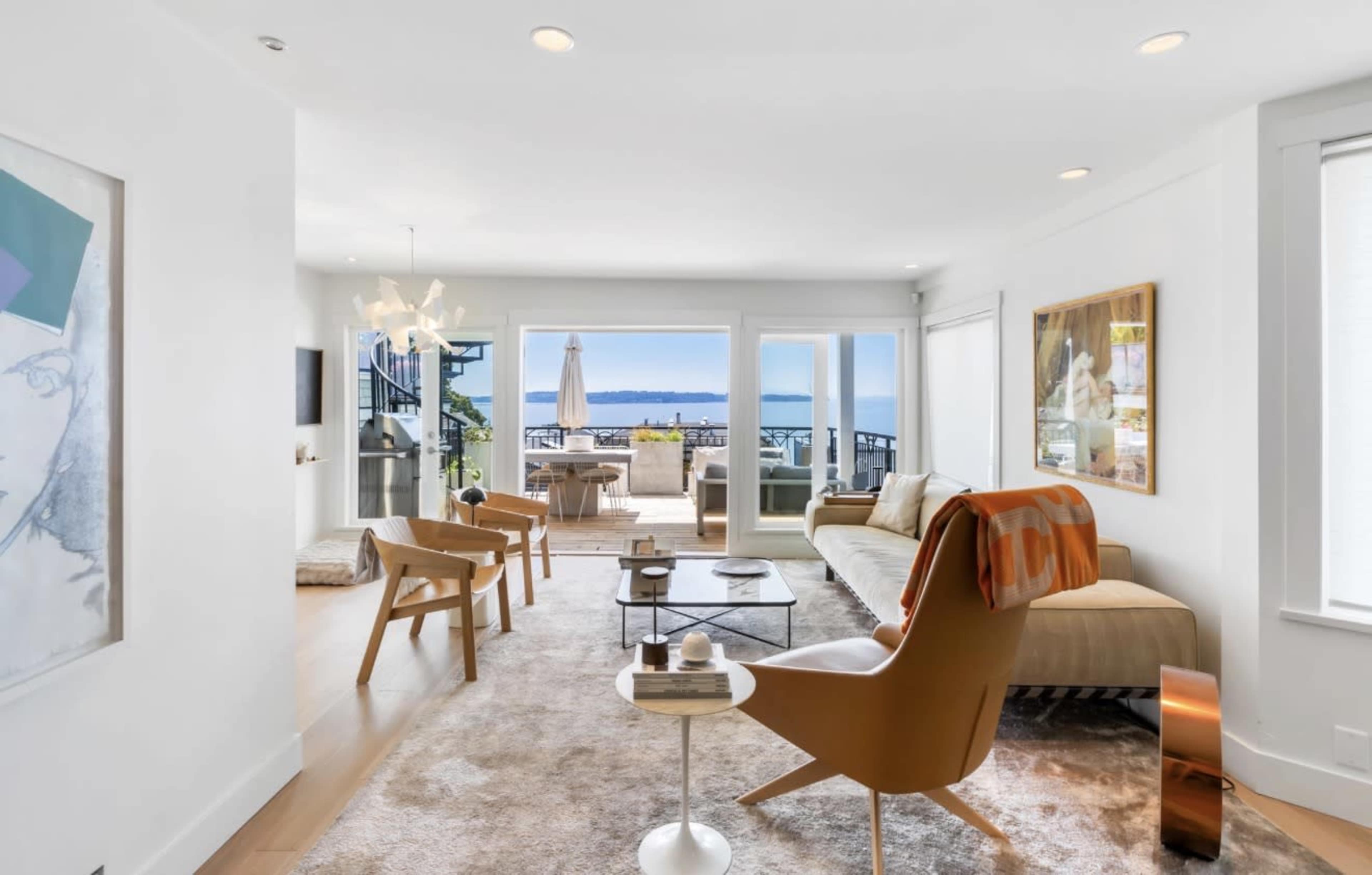 The image shows a modern living room with large windows overlooking the water, featuring stylish furniture and a light color scheme.