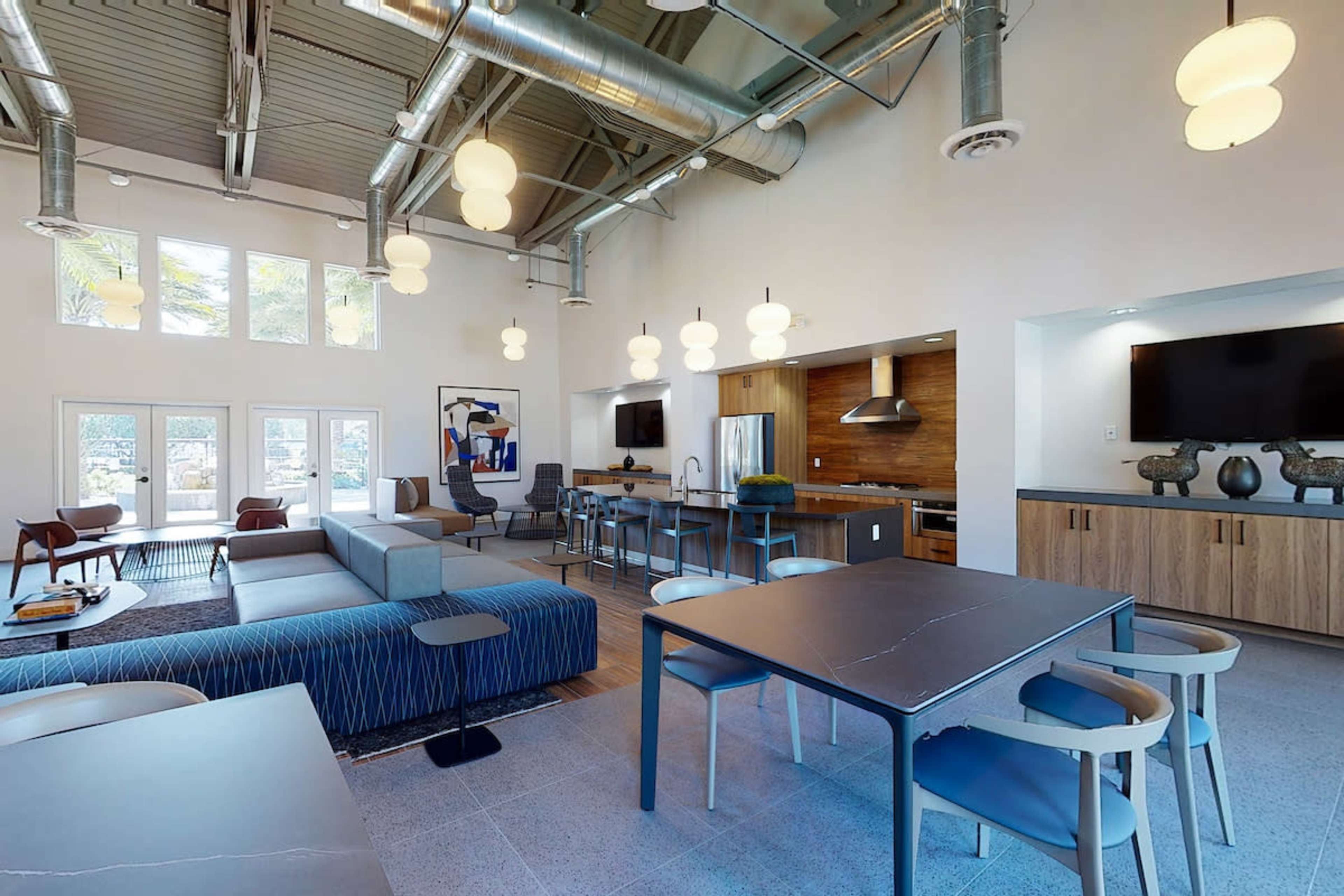 A modern communal living space with a kitchen area, dining table, and various seating arrangements under pendant lighting.