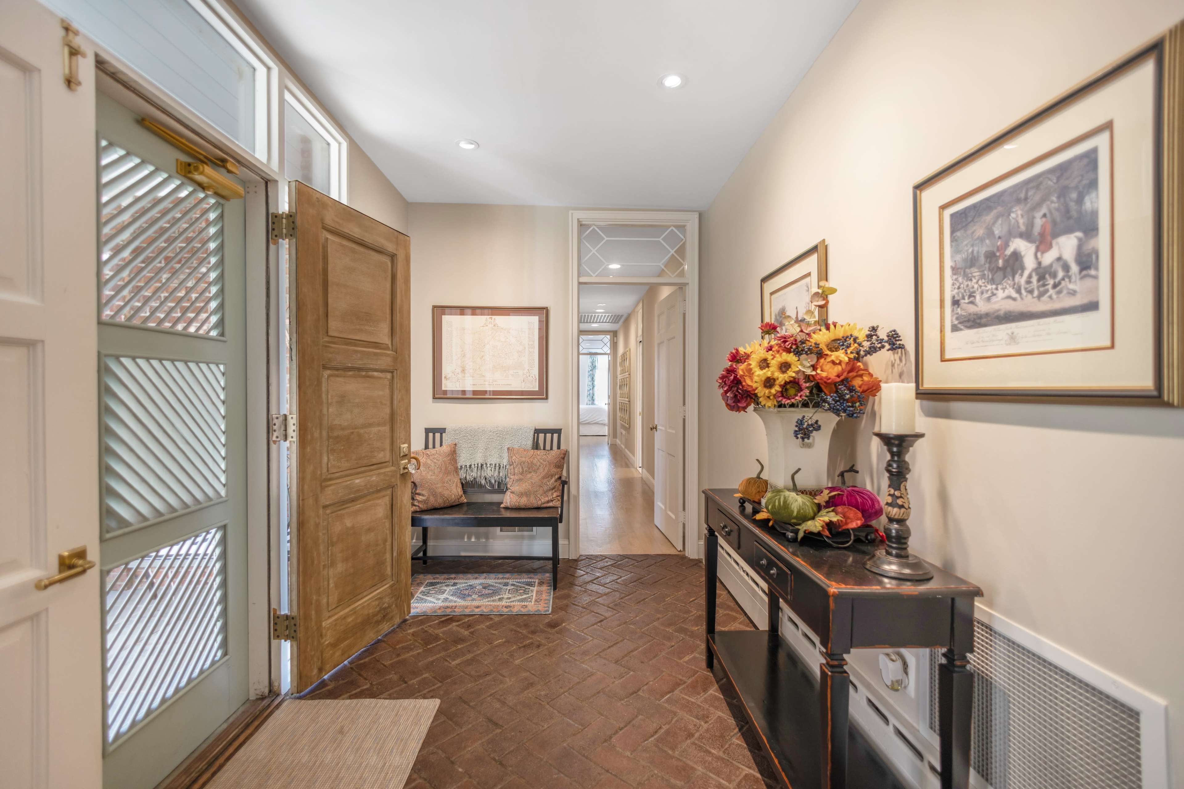 A hallway with a wooden door, a small seating area, decorative flowers on a console table, and an entrance leading to a corridor.