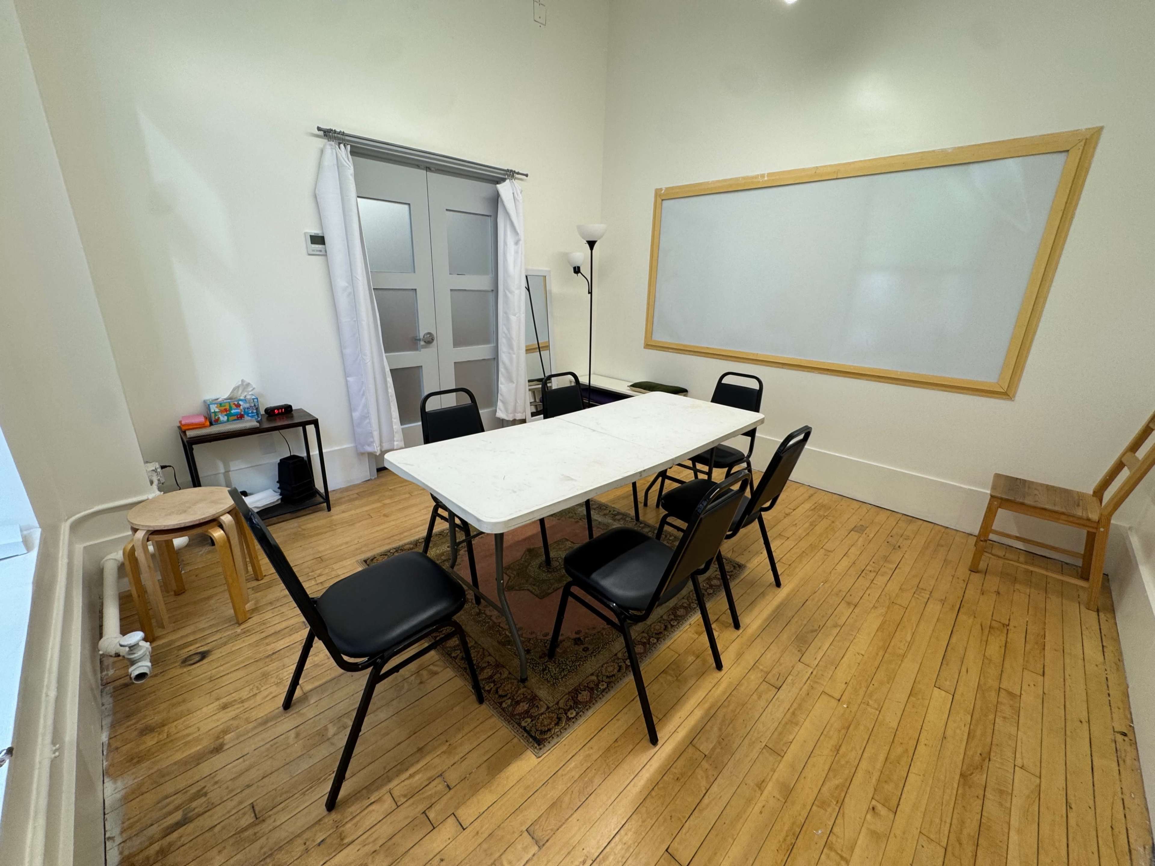 The image shows a simple meeting room with a rectangular table surrounded by black chairs, a floor lamp, a chalkboard, and a small stool.