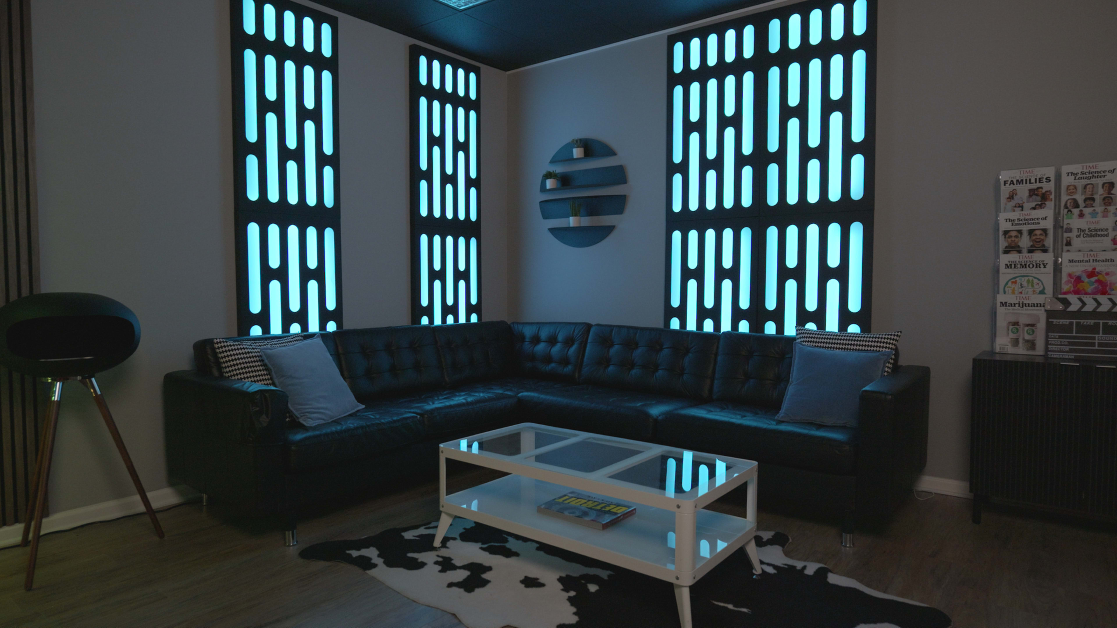 The image depicts a modern living room featuring a black sectional sofa, a glass coffee table, and illuminated wall panels with a blue design.