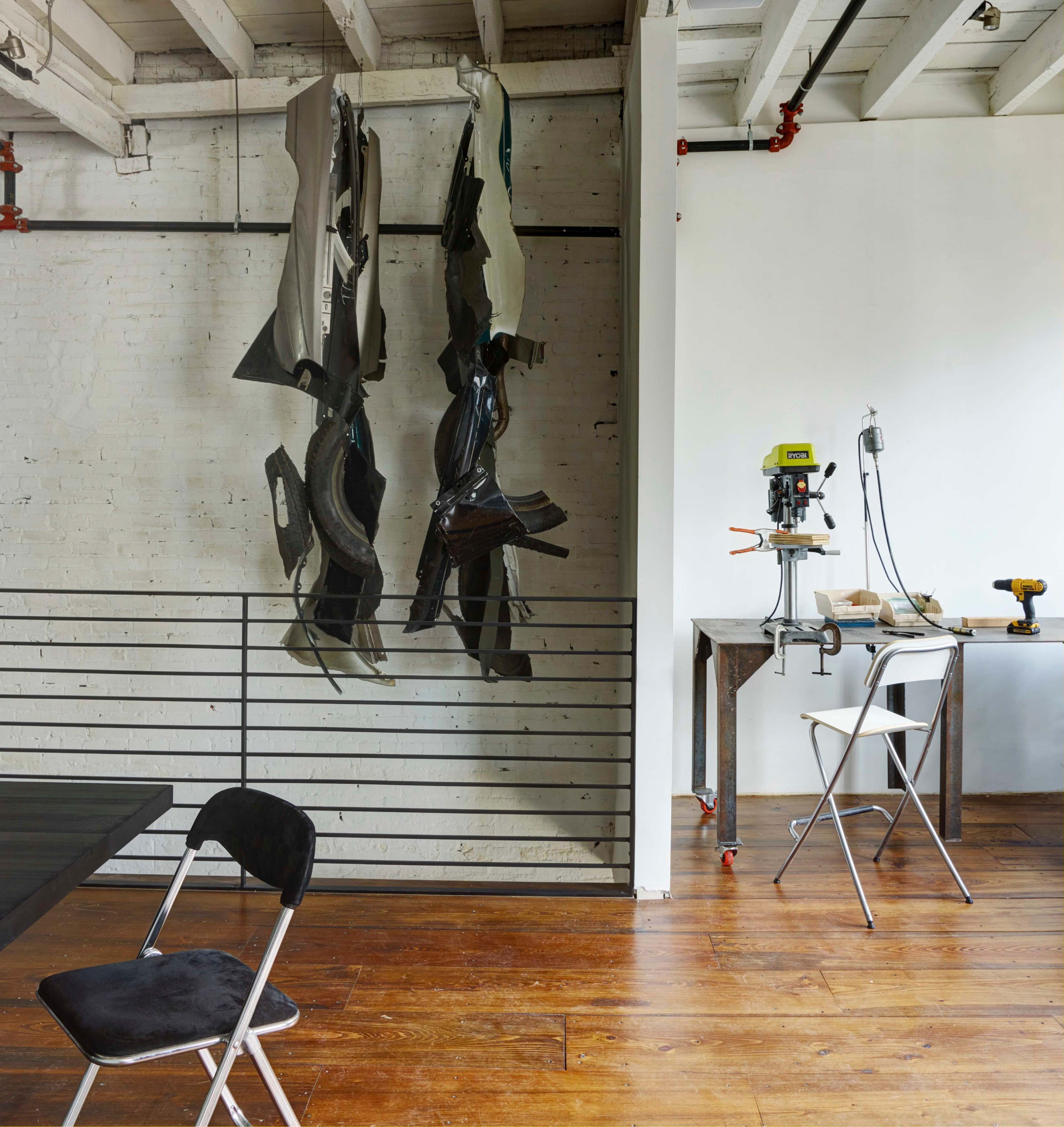 The image shows a modern workspace with hanging sculptural pieces on the wall and a workbench equipped with tools and chairs.
