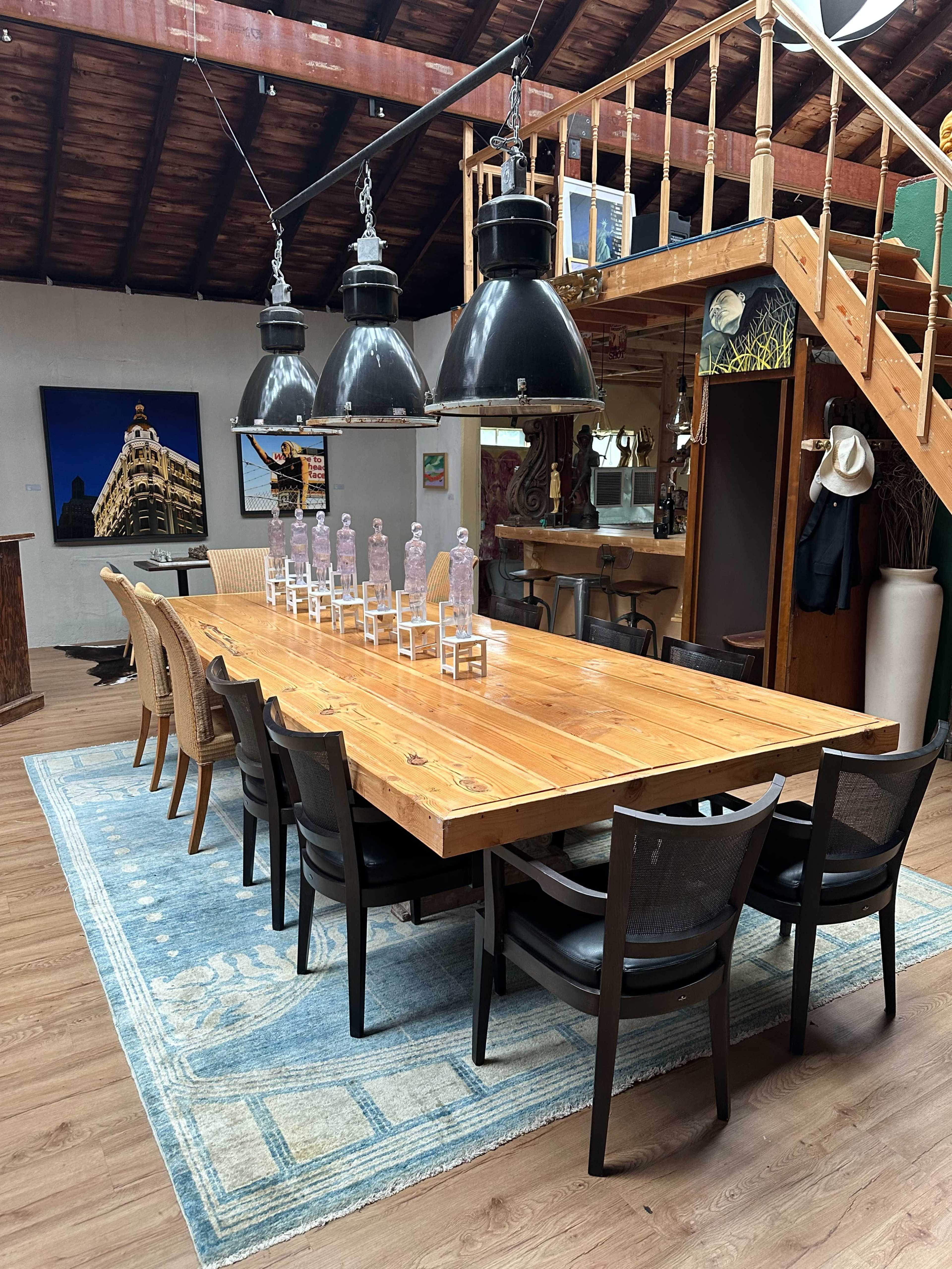 A long wooden table with black chairs is set in a spacious room, illuminated by large pendant lights, with a staircase and artwork visible in the background.