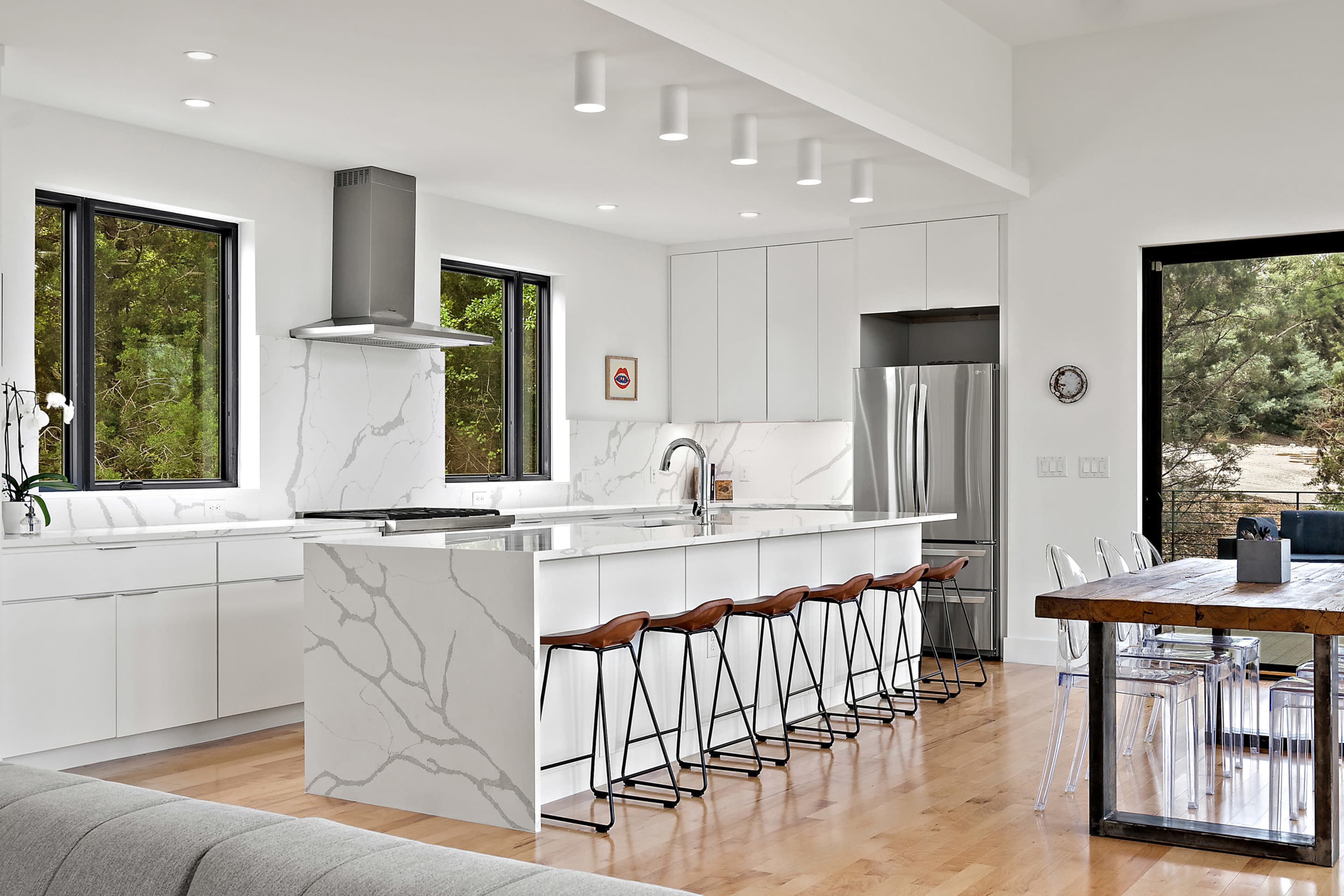 A modern kitchen features white cabinets, a large island with a marble pattern, and bar stools, with large windows providing natural light.