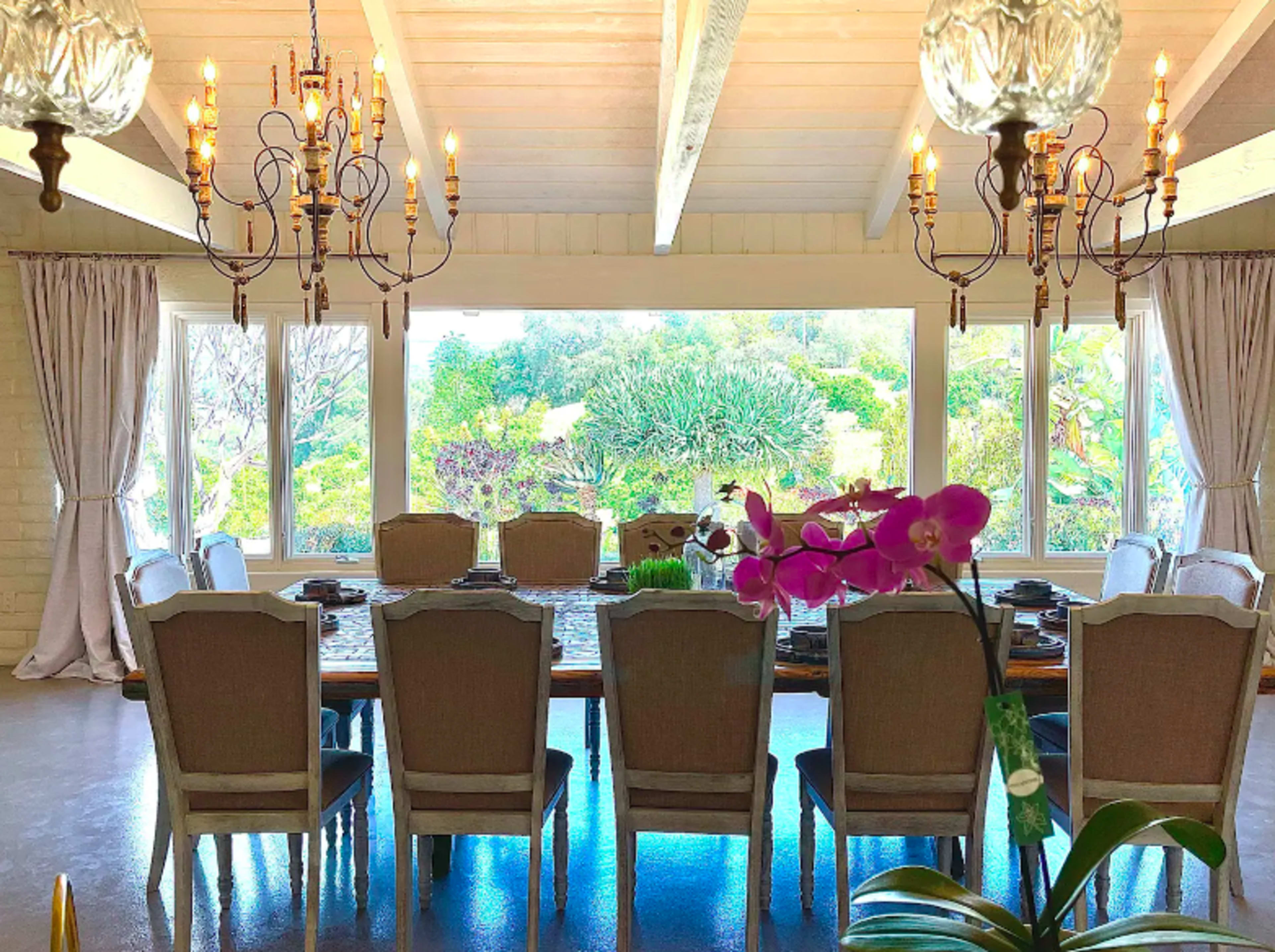 A spacious dining room features a long table surrounded by chairs, with large windows showcasing a lush garden view.