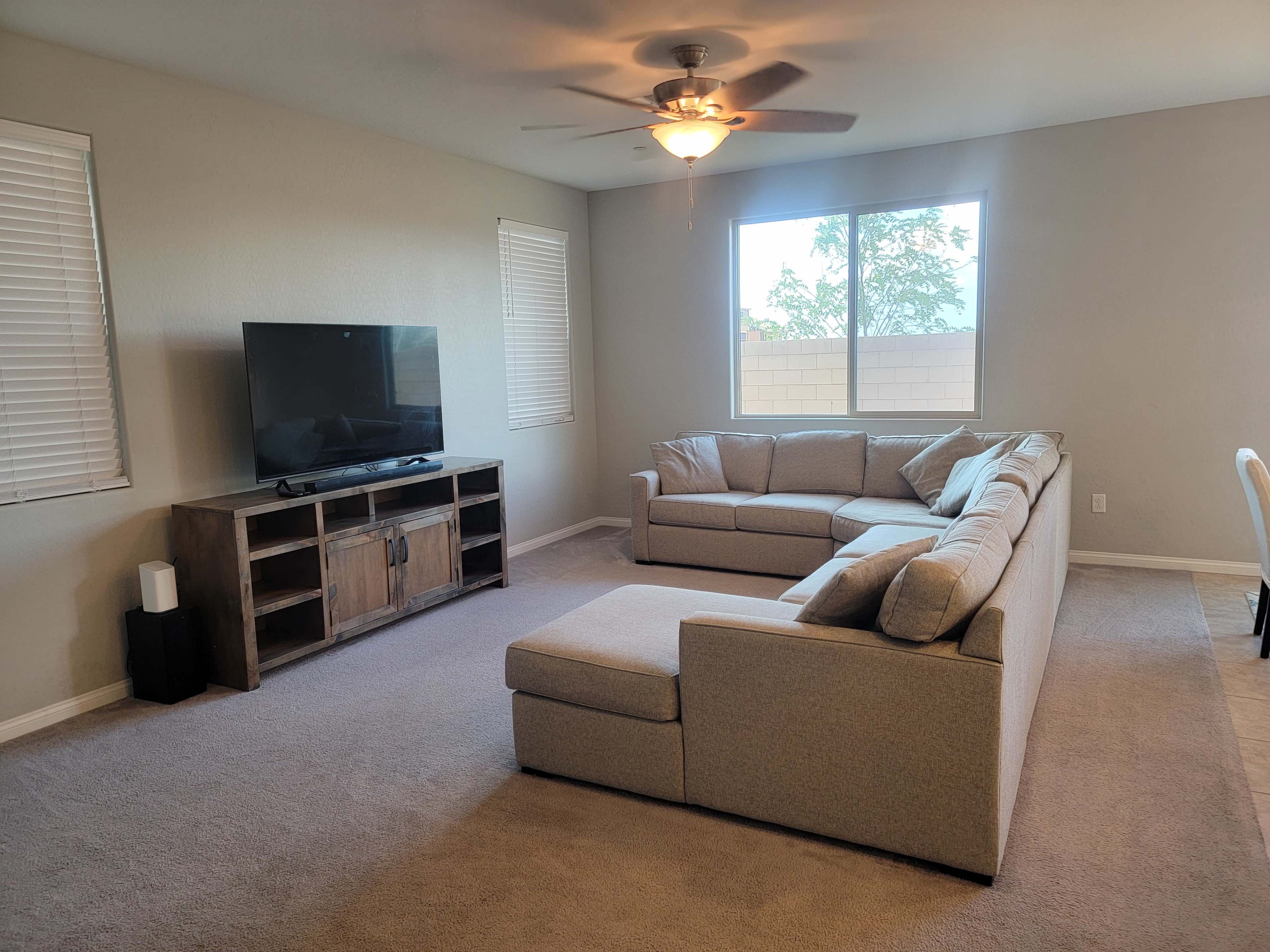 The living room features a large sectional sofa, a television on a wooden stand, and two windows letting in natural light.