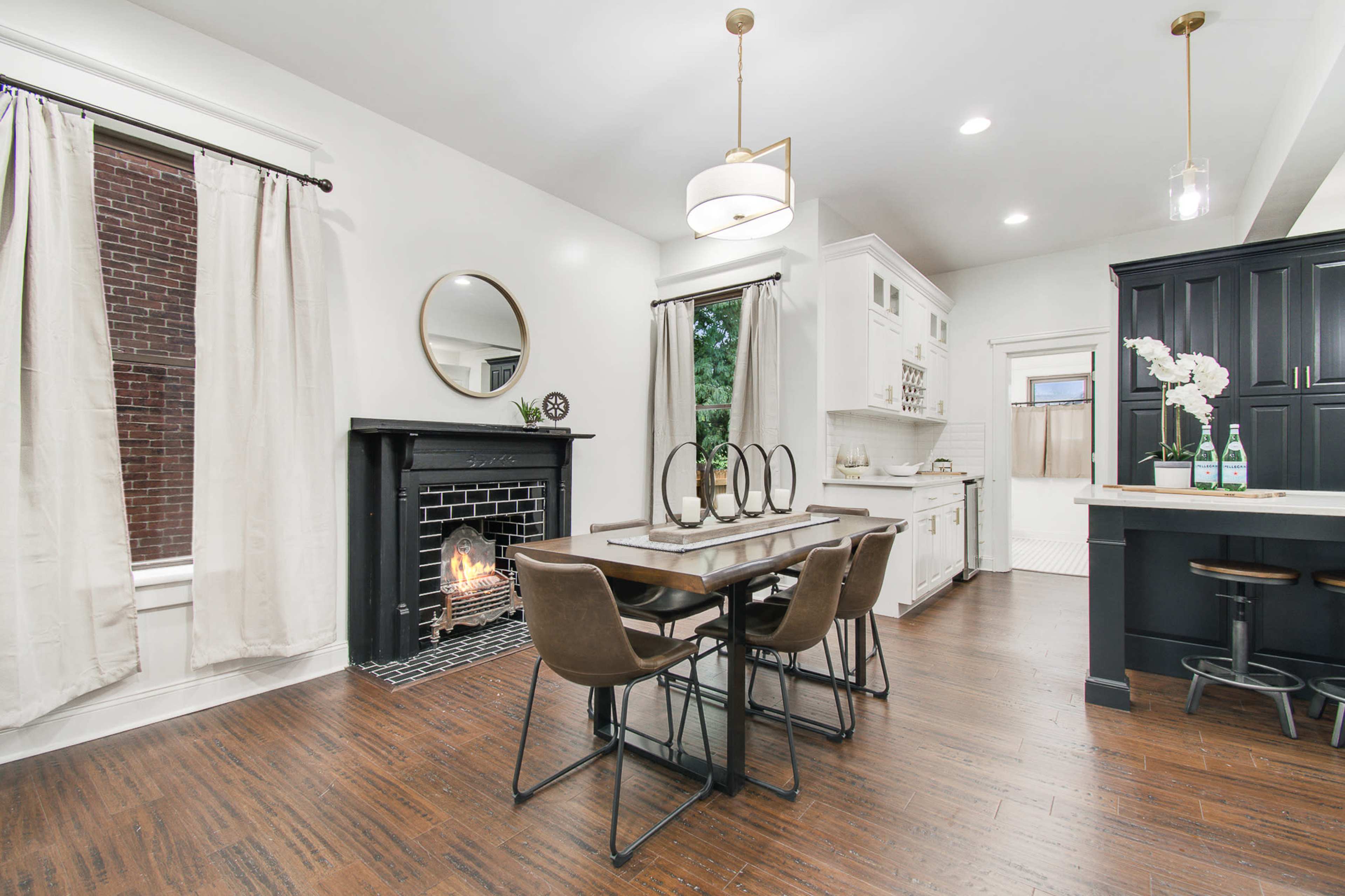 A dining area features a black fireplace, a wooden table with four chairs, and white walls adorned with light curtains.