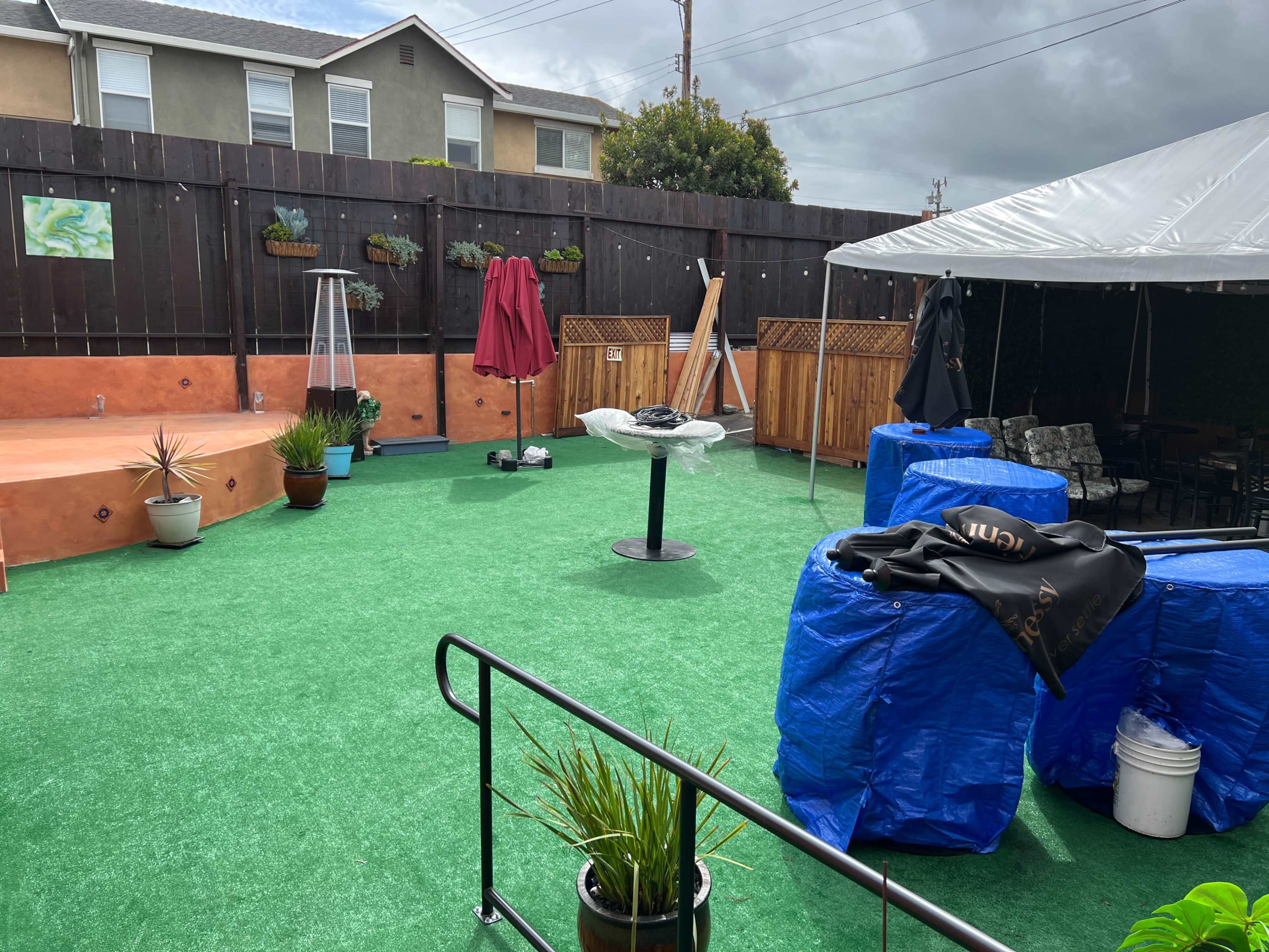 The image shows an outdoor area with artificial turf, a large tent, and various plants and furniture, with partially covered tables and a few decorative items.