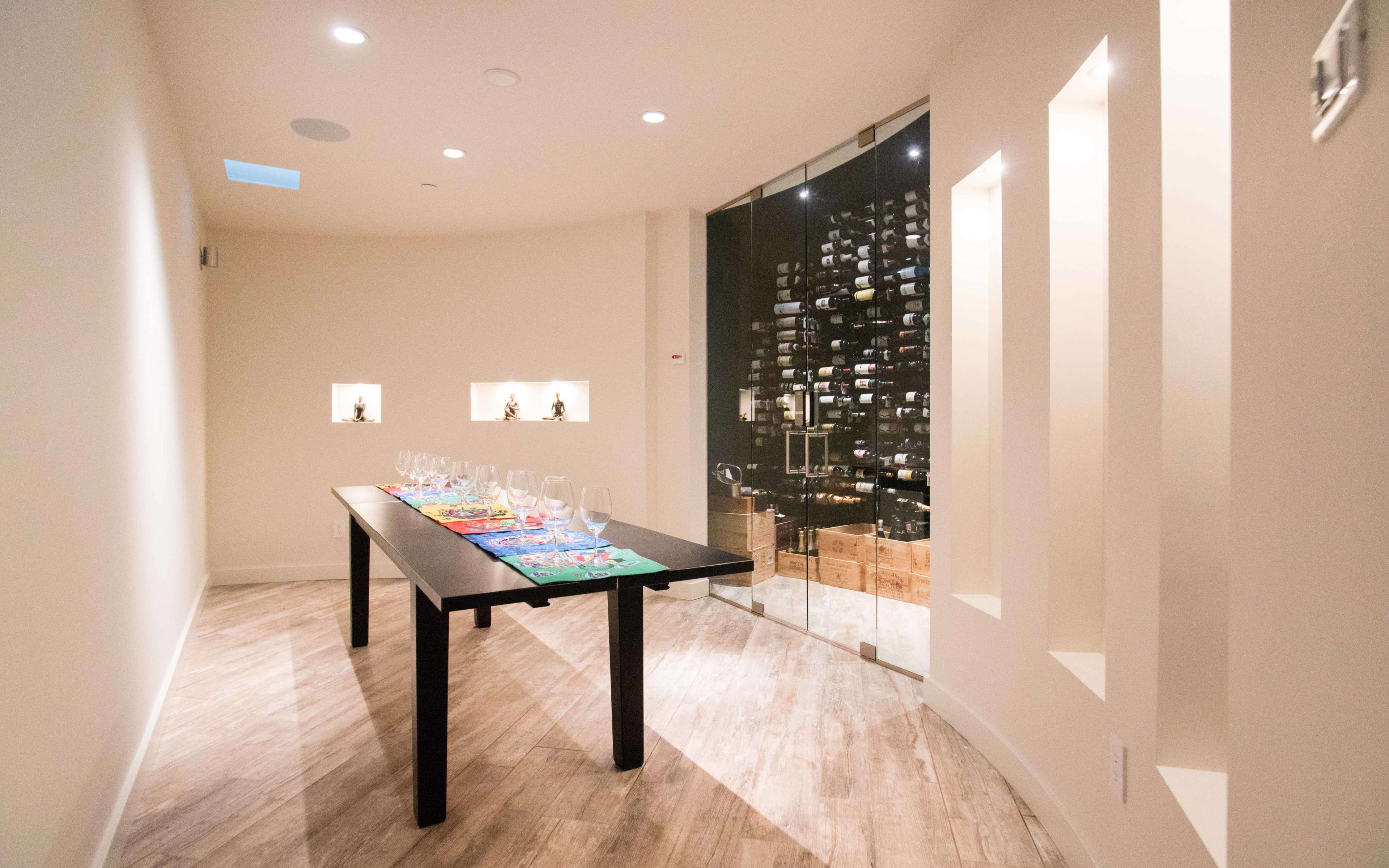 A sleek wine tasting room features a long black table set with glasses, adjacent to a glass wall displaying a wine cellar.