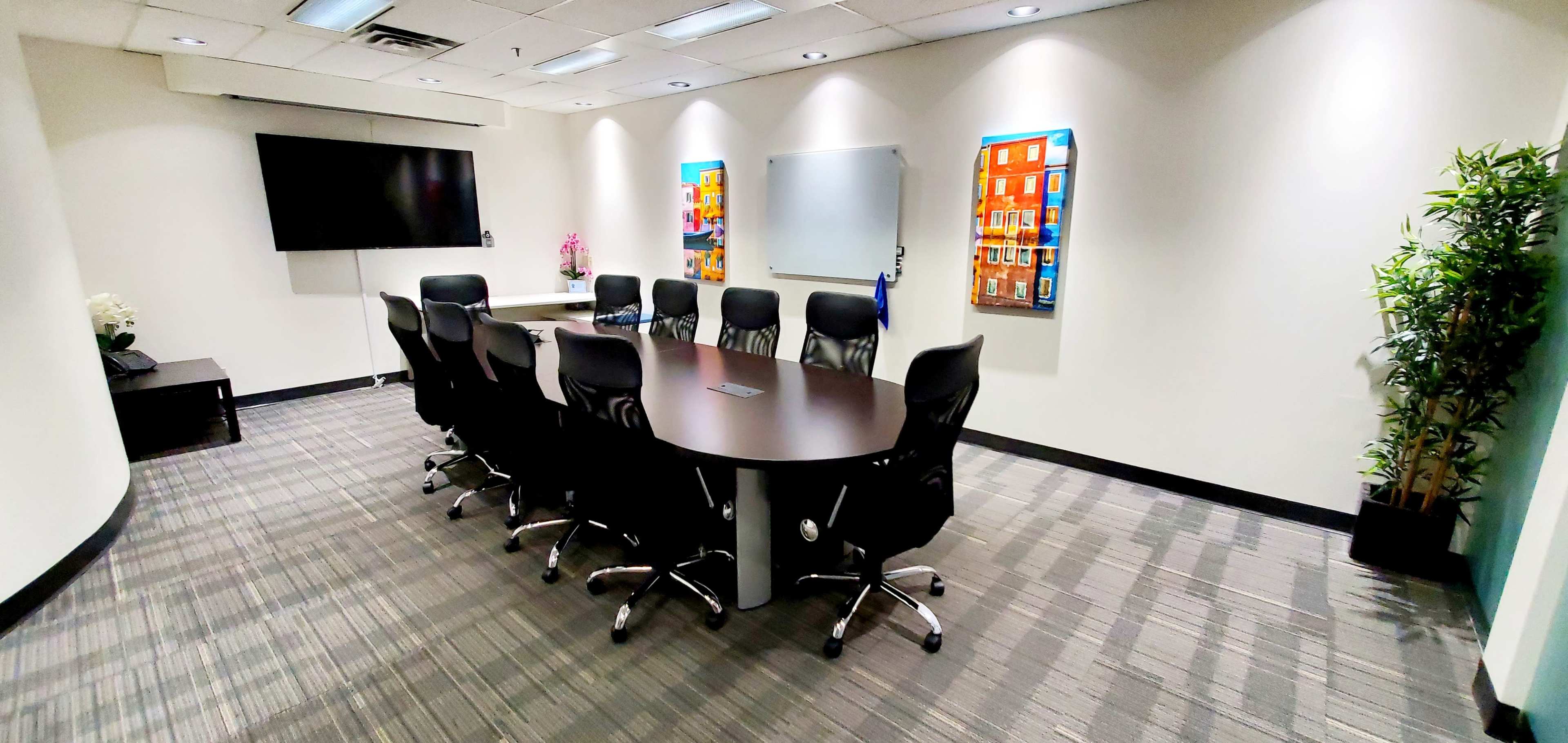 A conference room features a large oval table surrounded by nine black chairs, with a television screen and whiteboard on one wall and colorful artwork on another.