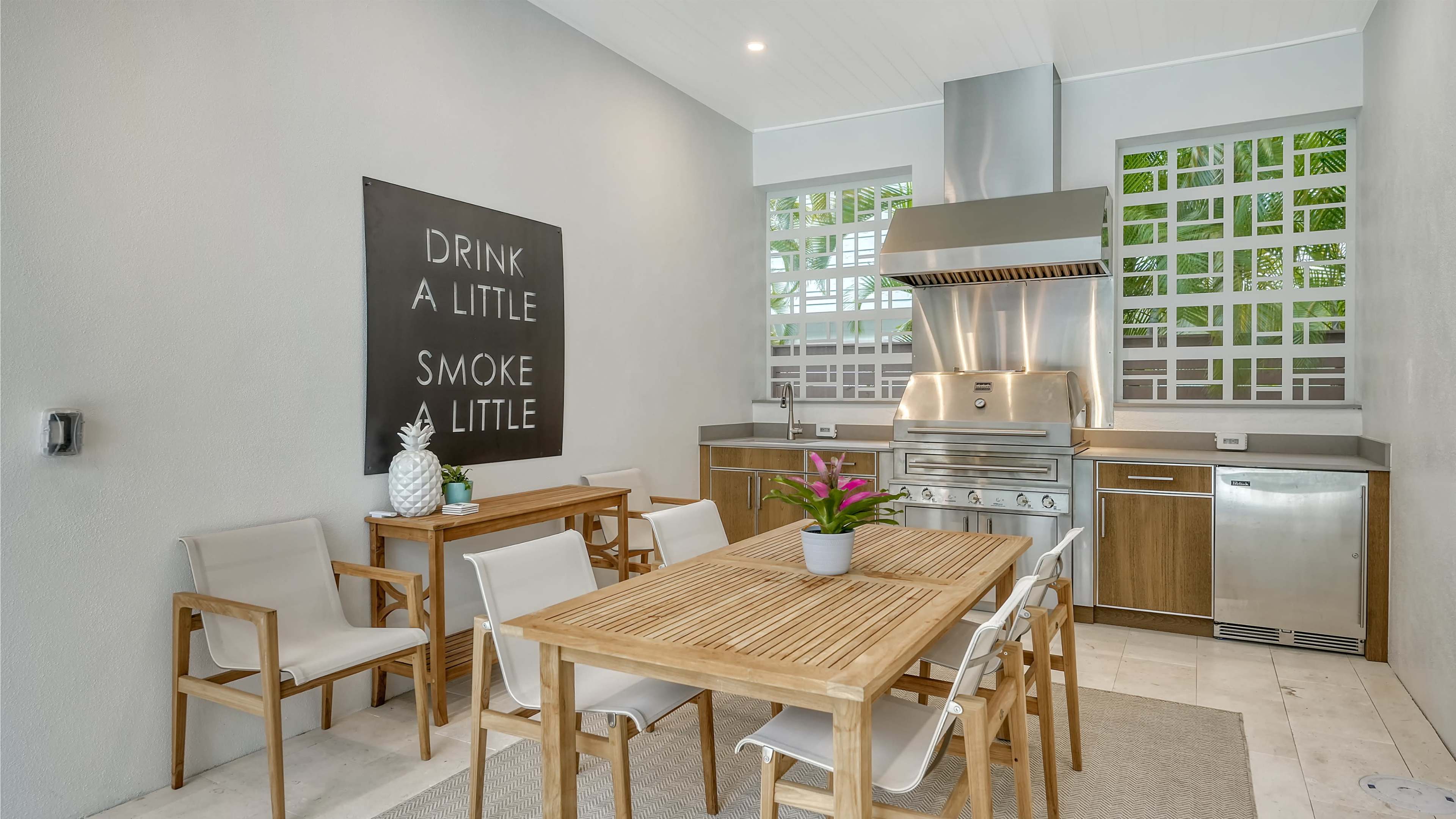 A modern outdoor kitchen area featuring a stainless steel grill, a wood dining table with eight chairs, and a large wall sign with the text "DRINK A LITTLE SMOKE A LITTLE."