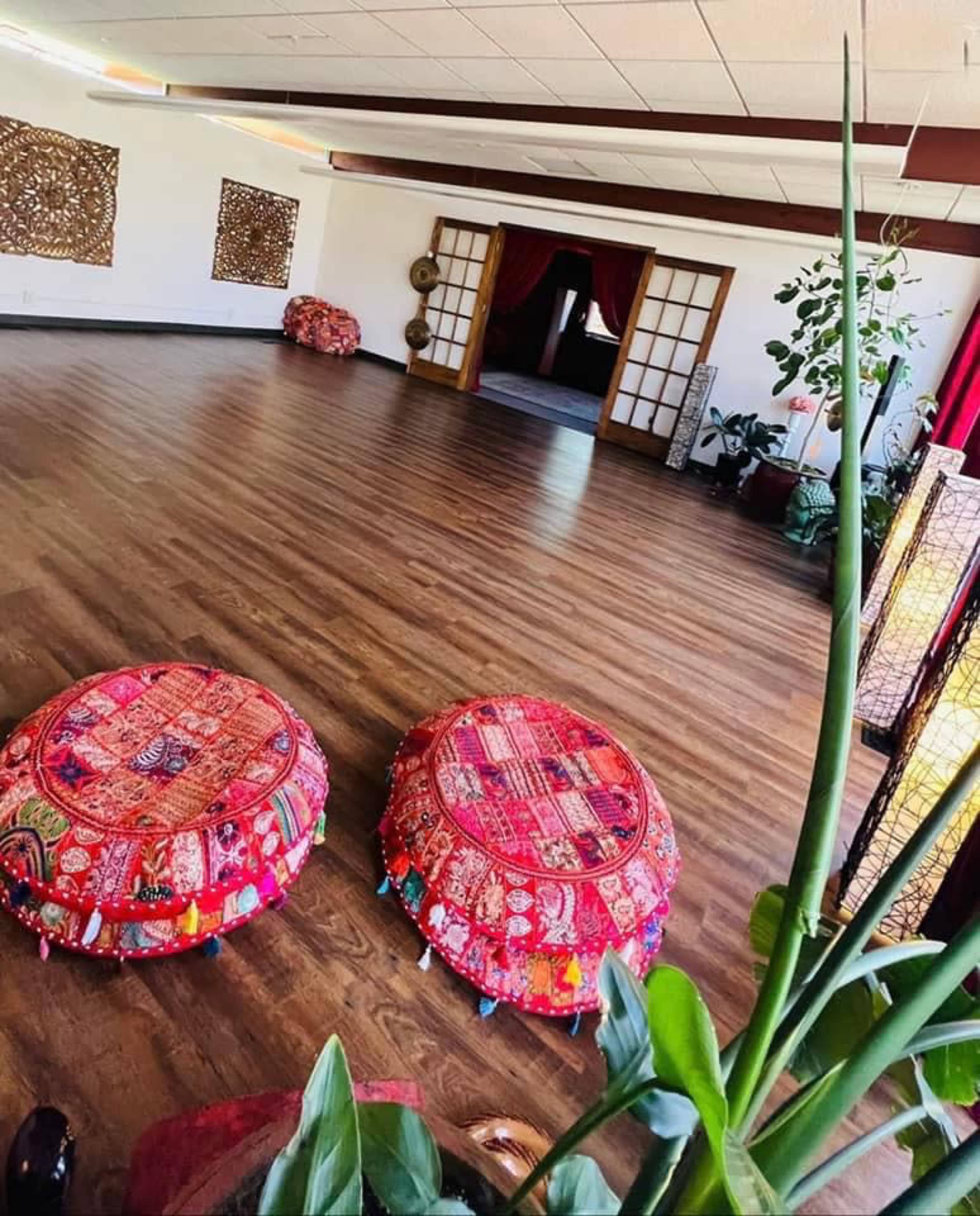 A spacious room with wooden flooring, adorned with decorative wall art, two vibrant floor cushions, and plants beside lanterns.