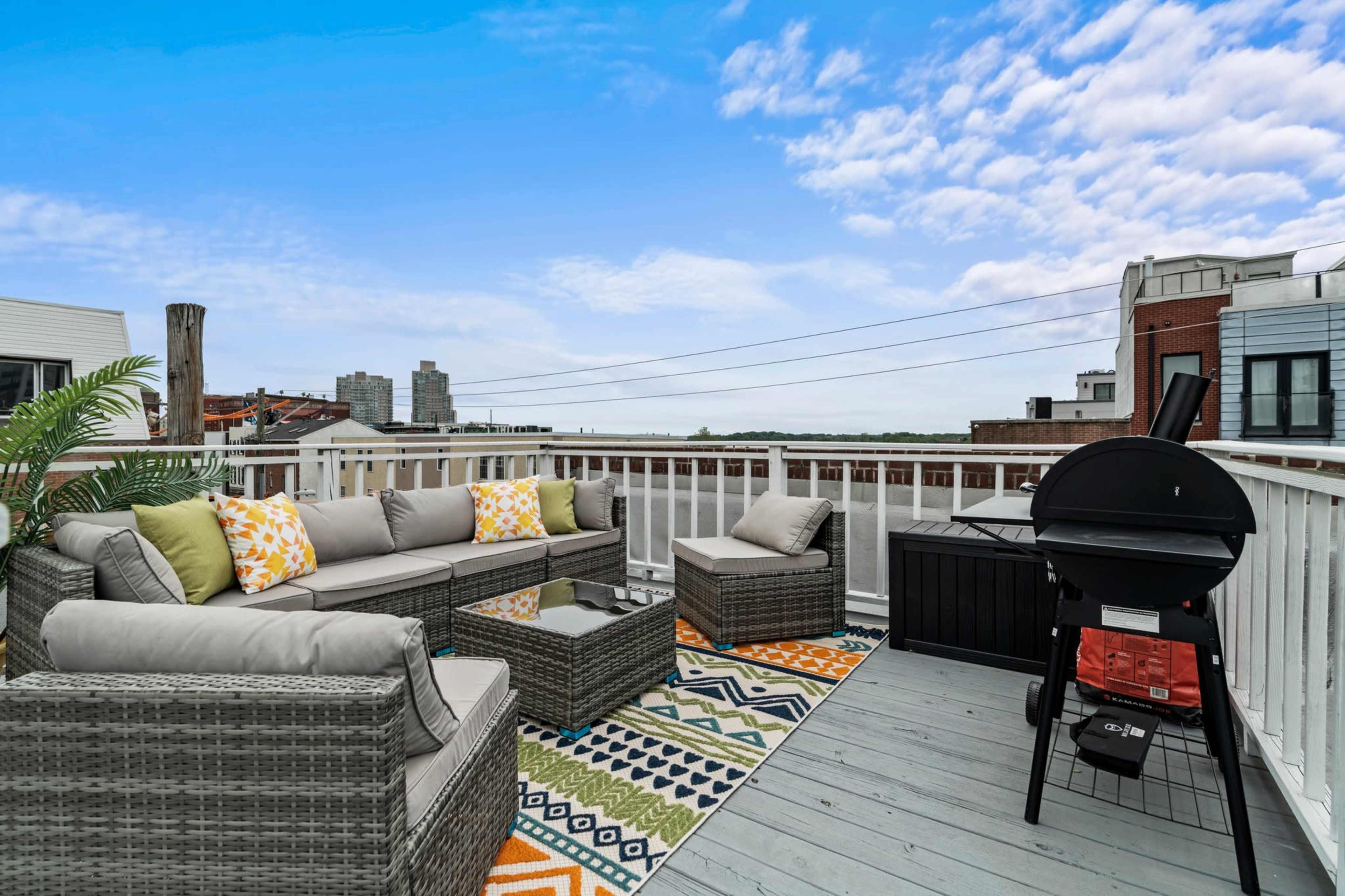 The image shows a rooftop patio with seating arranged around a table, a grill, and decorative cushions, overlooking a city skyline.