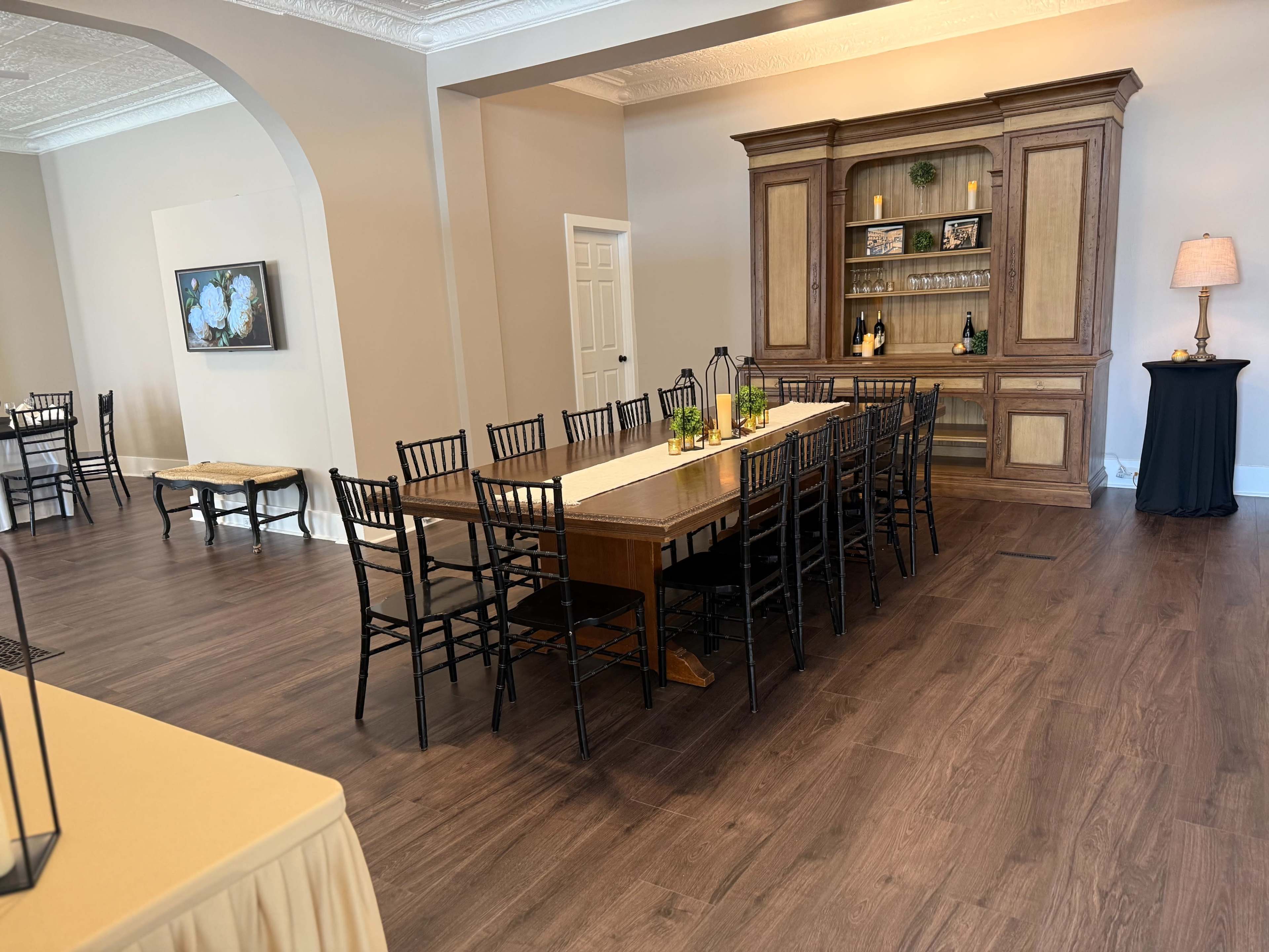 The image shows a spacious room with a long table set for dining, surrounded by black chairs, and a buffet-style cabinet against the wall.