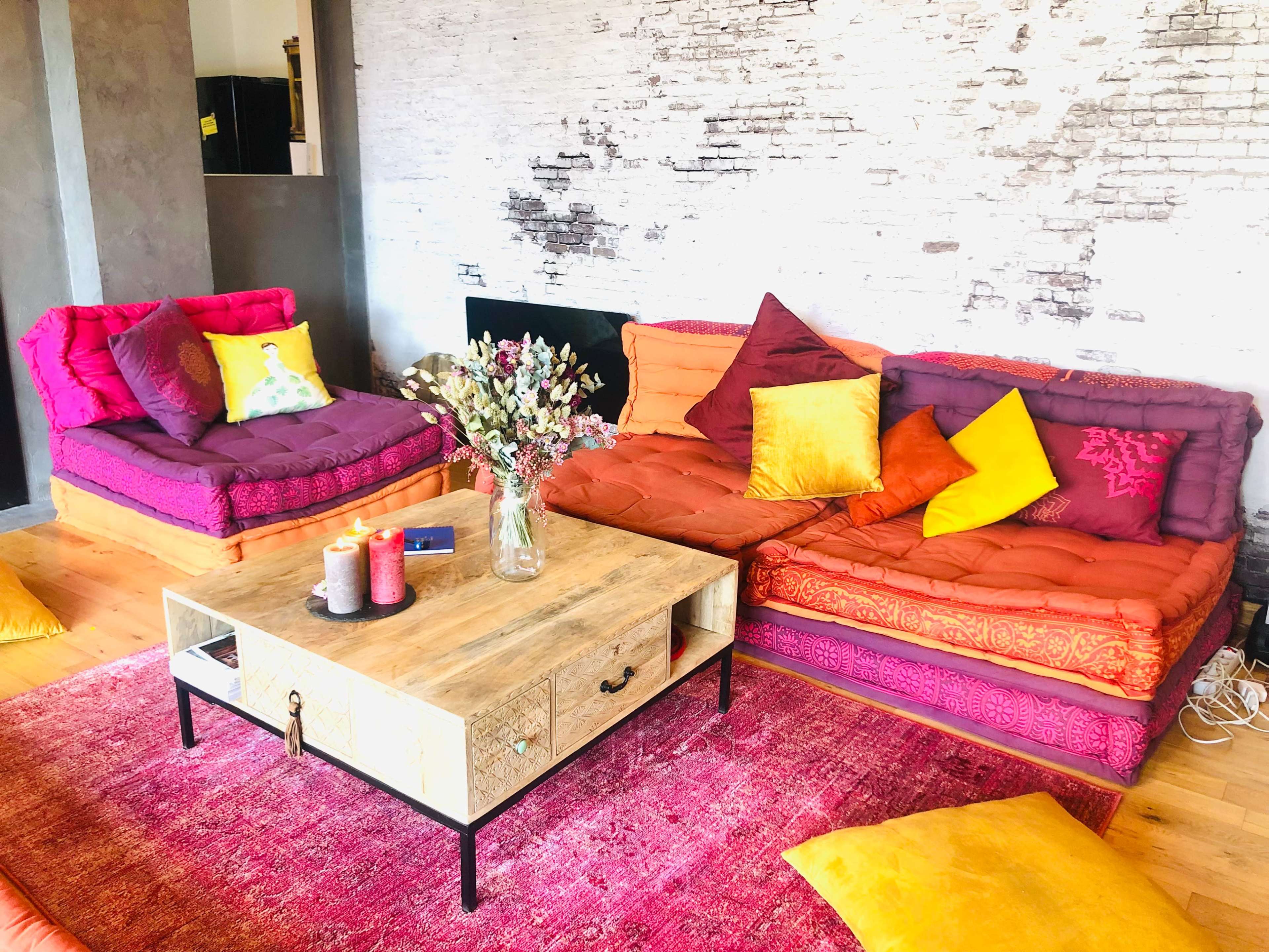 The image shows a brightly colored living room with two couches, a wooden coffee table, and a vase of flowers on a vibrant rug.